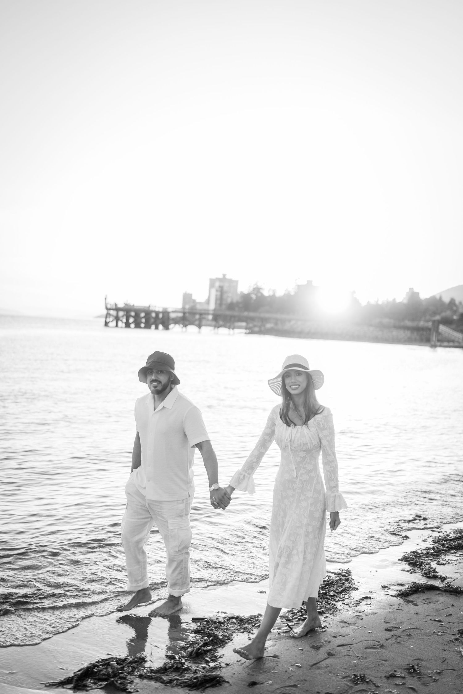 A couple in white outfits walks along a misty beach shoreline at sunset creating a romantic silhouette.