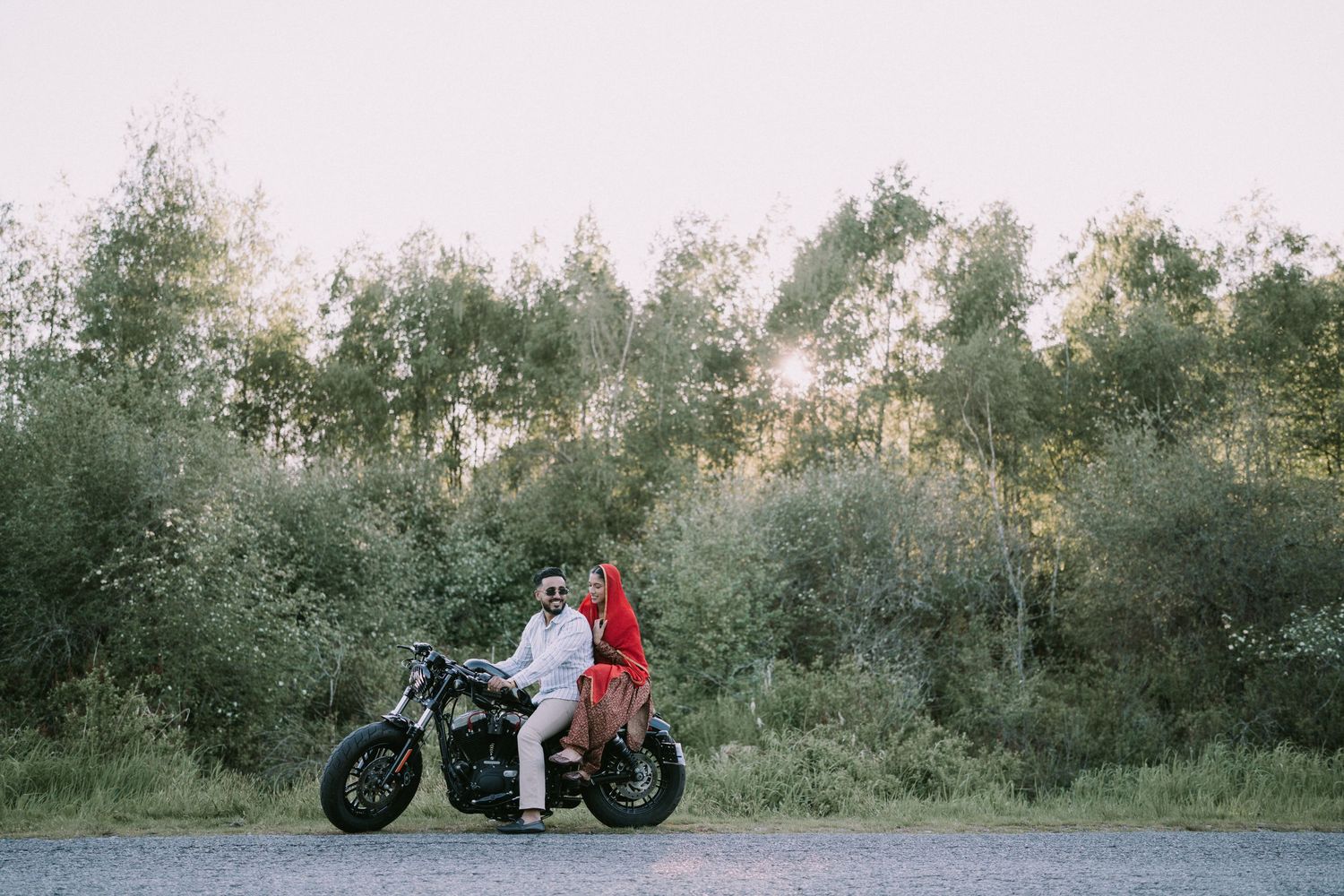 A couple sits together on a motorcycle parked along a tree-lined path at sunset.