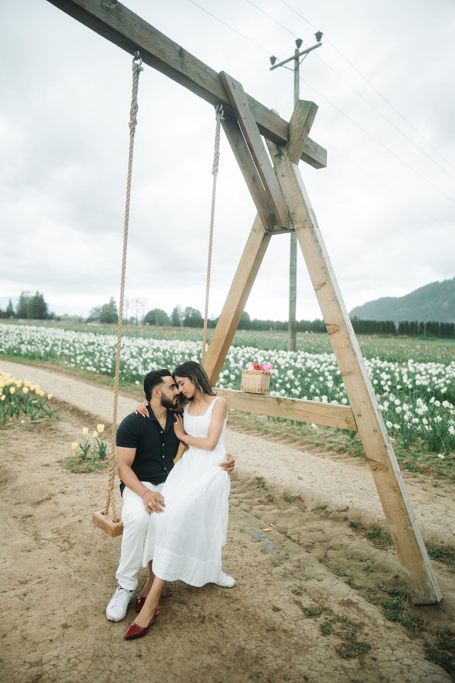 Couple sits together on wooden swing in field of white tulips on overcast day.