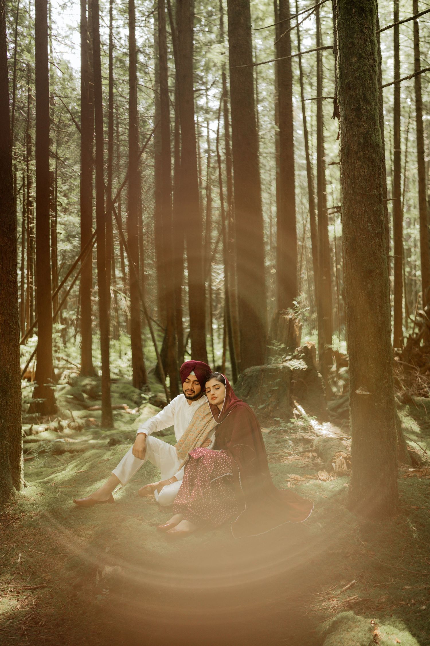 Sikh couple sitting together in a sunlit forest, man in white kurta and purple turban, woman in burgundy salwar kameez.