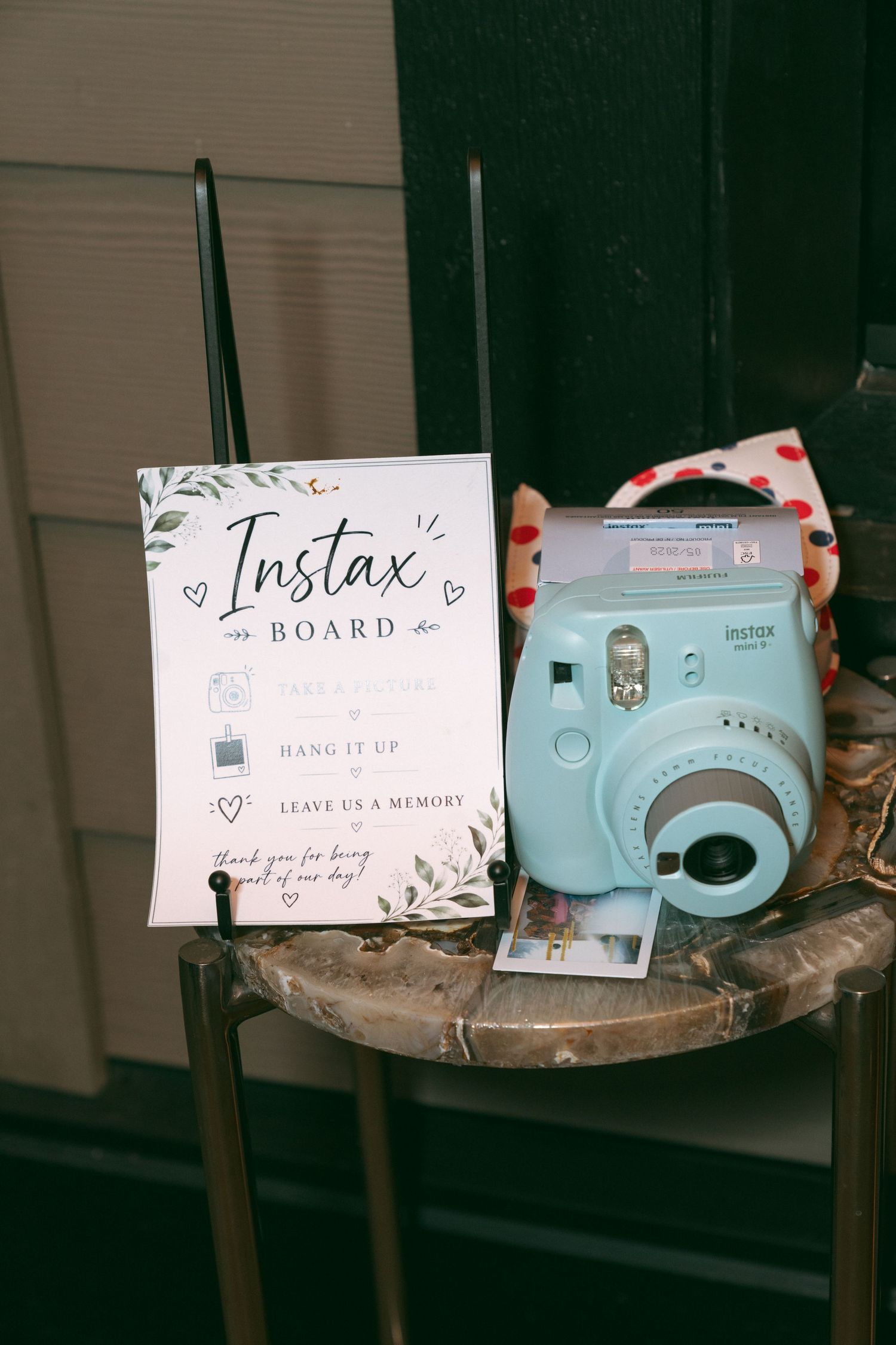 Fujifilm Instax Mini camera beside an Instax Board sign with greenery decor at a wedding reception.