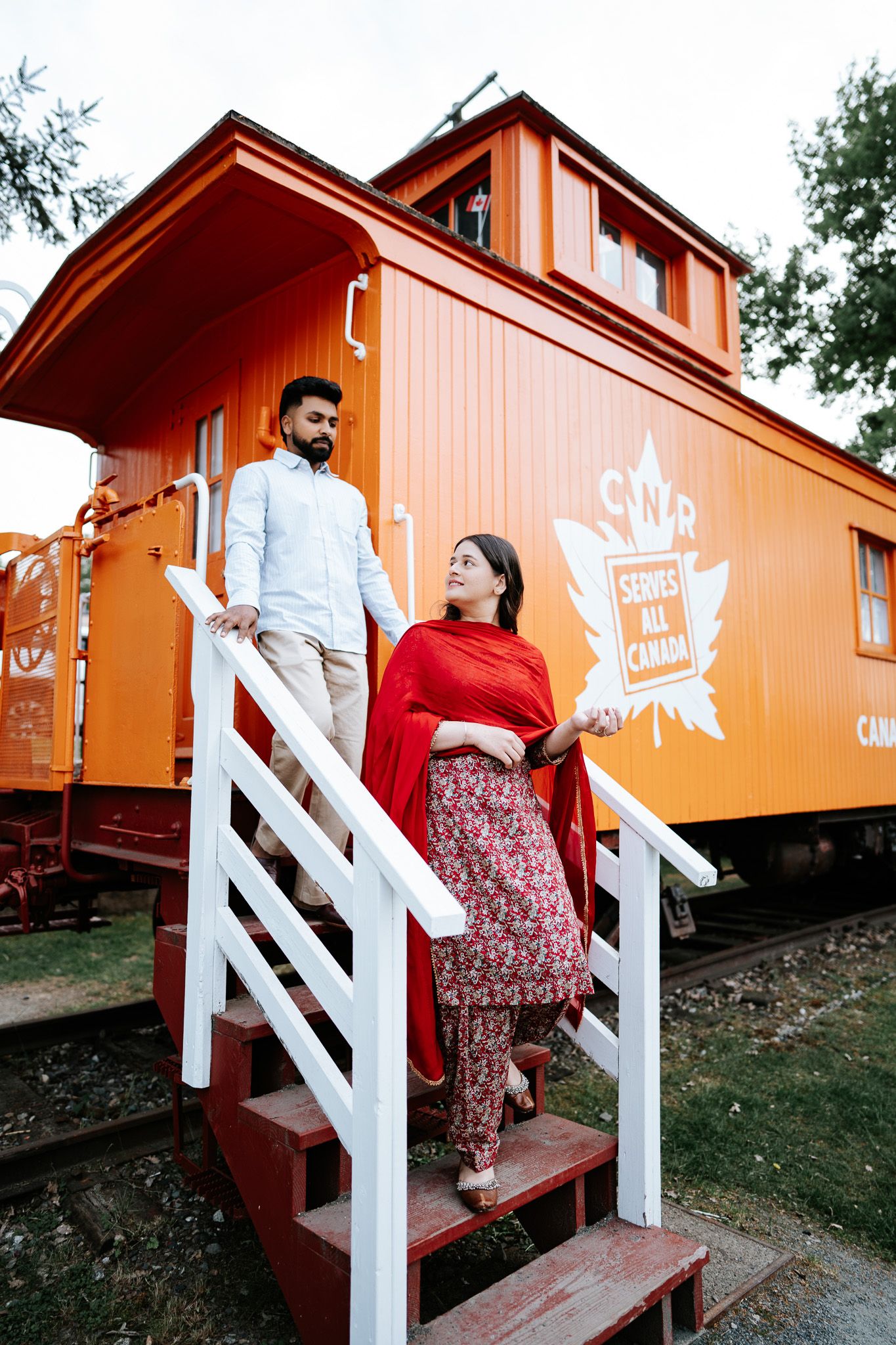 Couple in traditional South Asian attire on red stairs beside a vintage orange Canadian railway caboose.