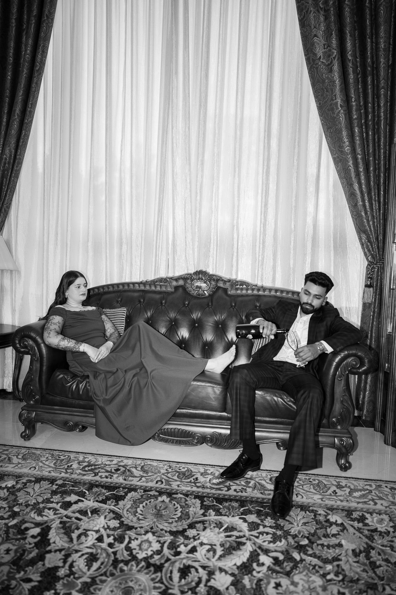 Sophisticated black and white portrait of a bearded man and tattooed woman in evening wear relaxing on ornate leather sofa.