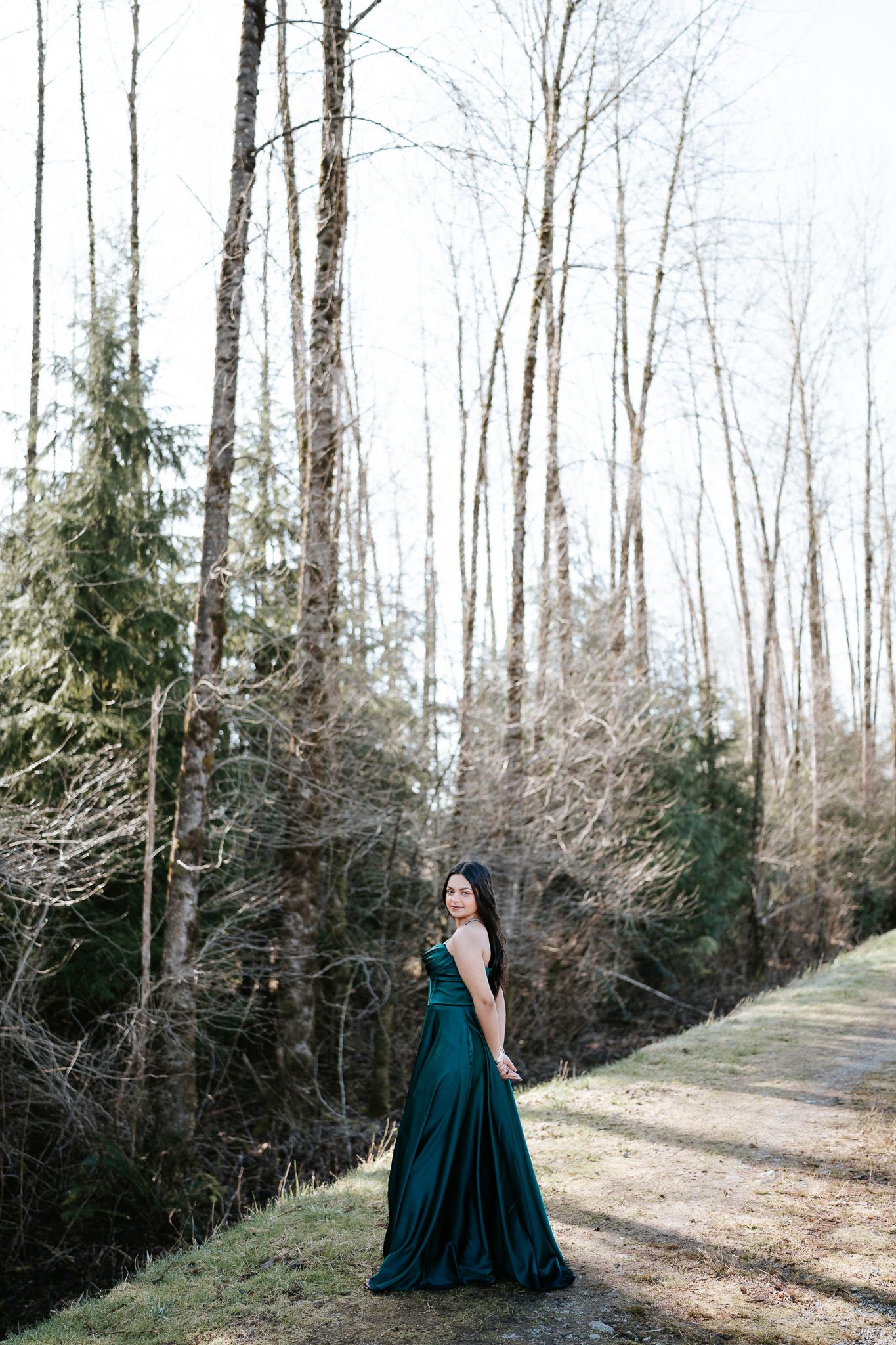A person in an elegant emerald green gown poses gracefully along a wooded path on a winter day.