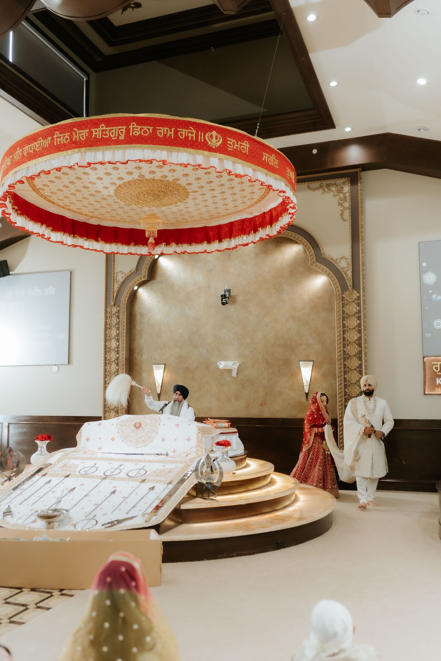 Sikh wedding ceremony inside a Gurdwara with ornate canopy, groom in white sherwani and bride in red lehenga.