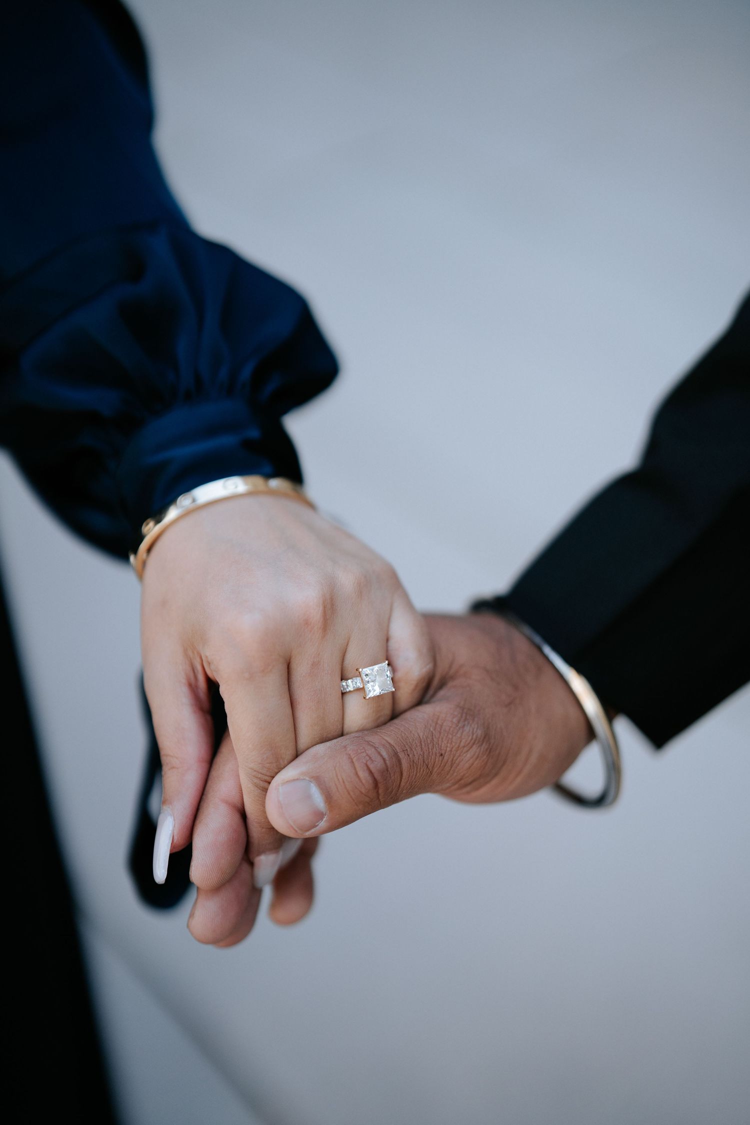 Two hands touching, one wearing a diamond engagement ring and gold bracelet, the other in a black suit sleeve with a gold cuff.