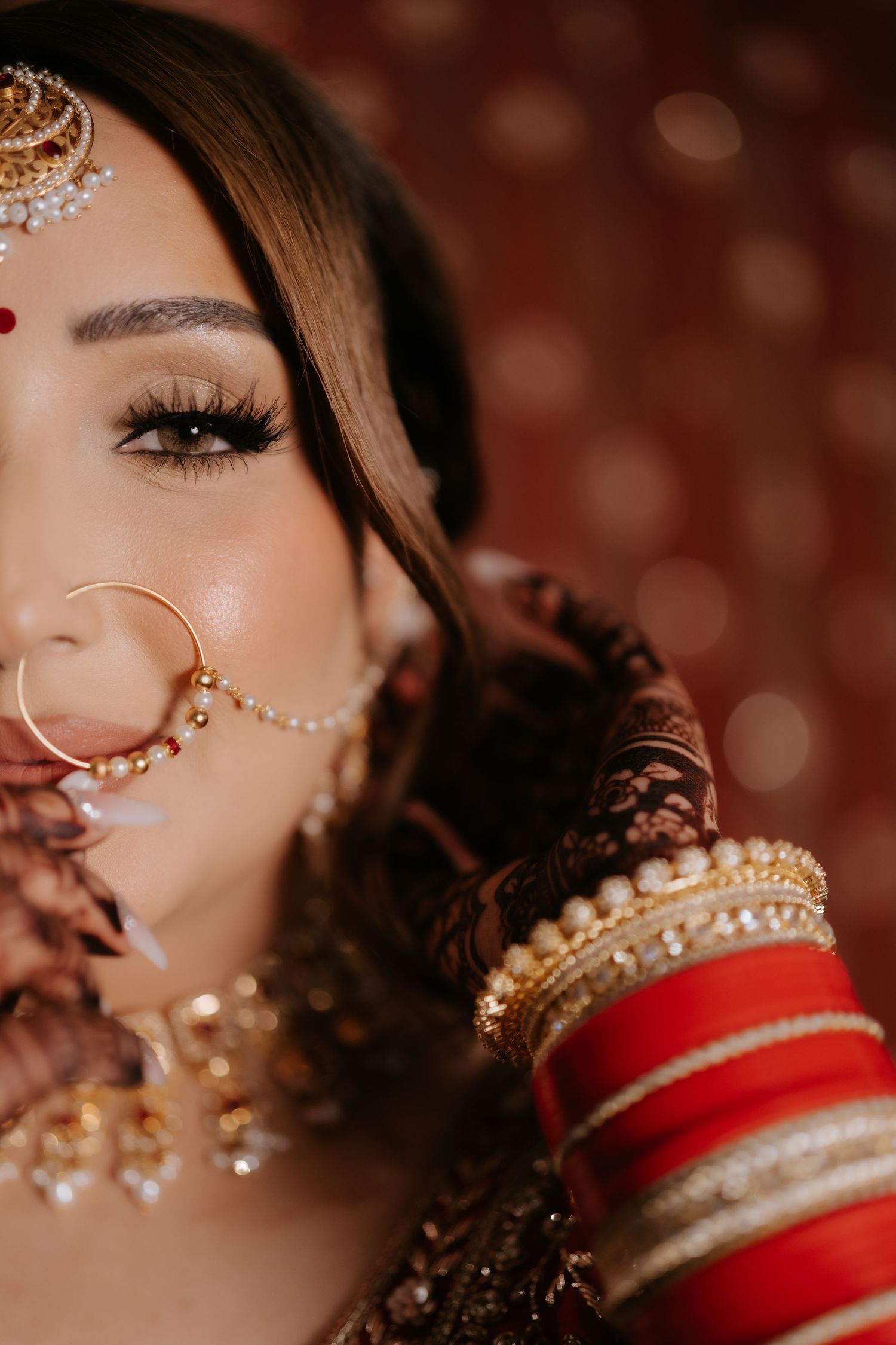 Close-up of Indian bride wearing traditional gold nath nose ring, red bangles, and ornate jewelry on wedding day.