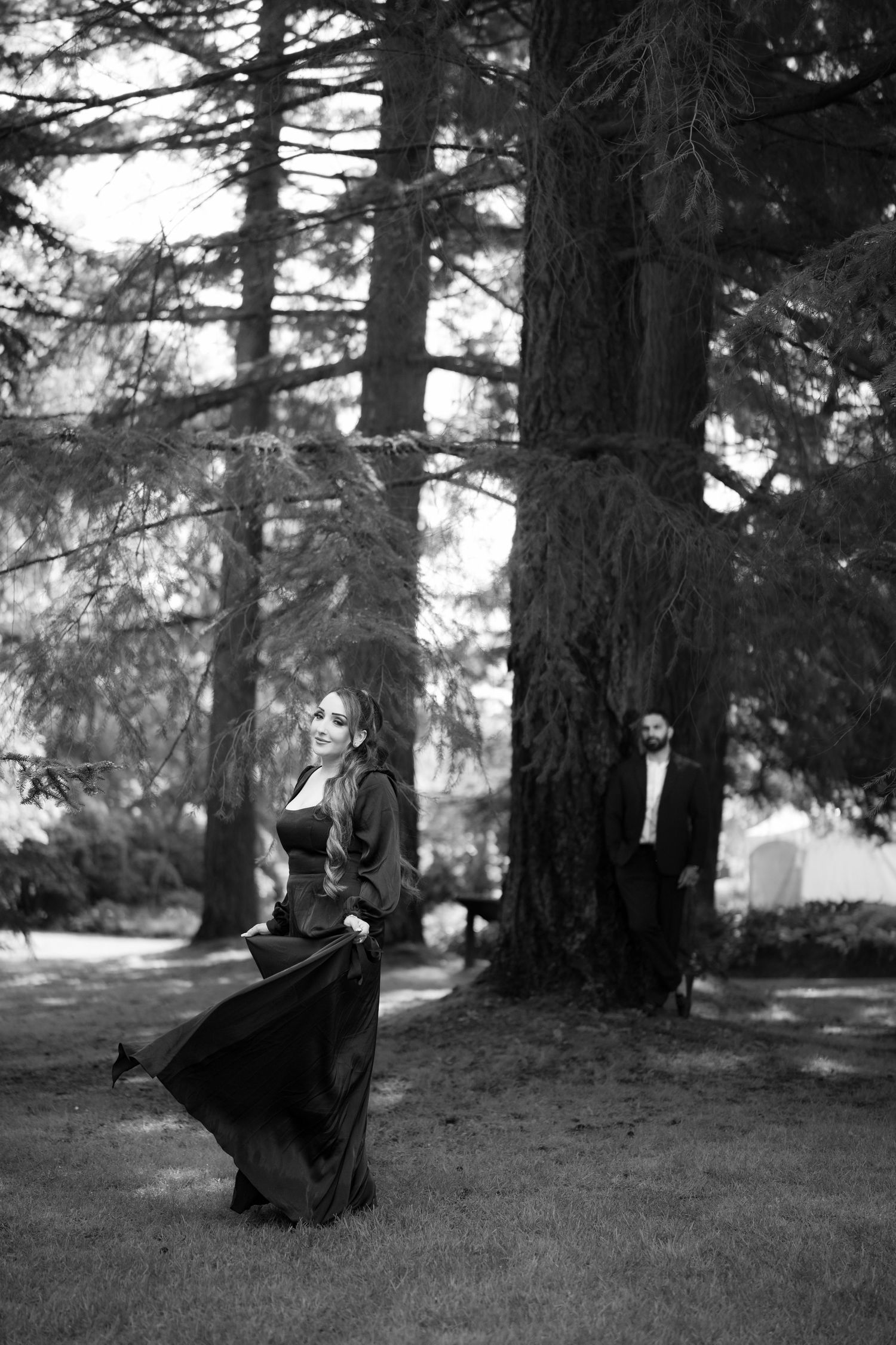 Black and white photo of a woman in flowing dark gown twirling outdoors as a man watches from behind tall trees.