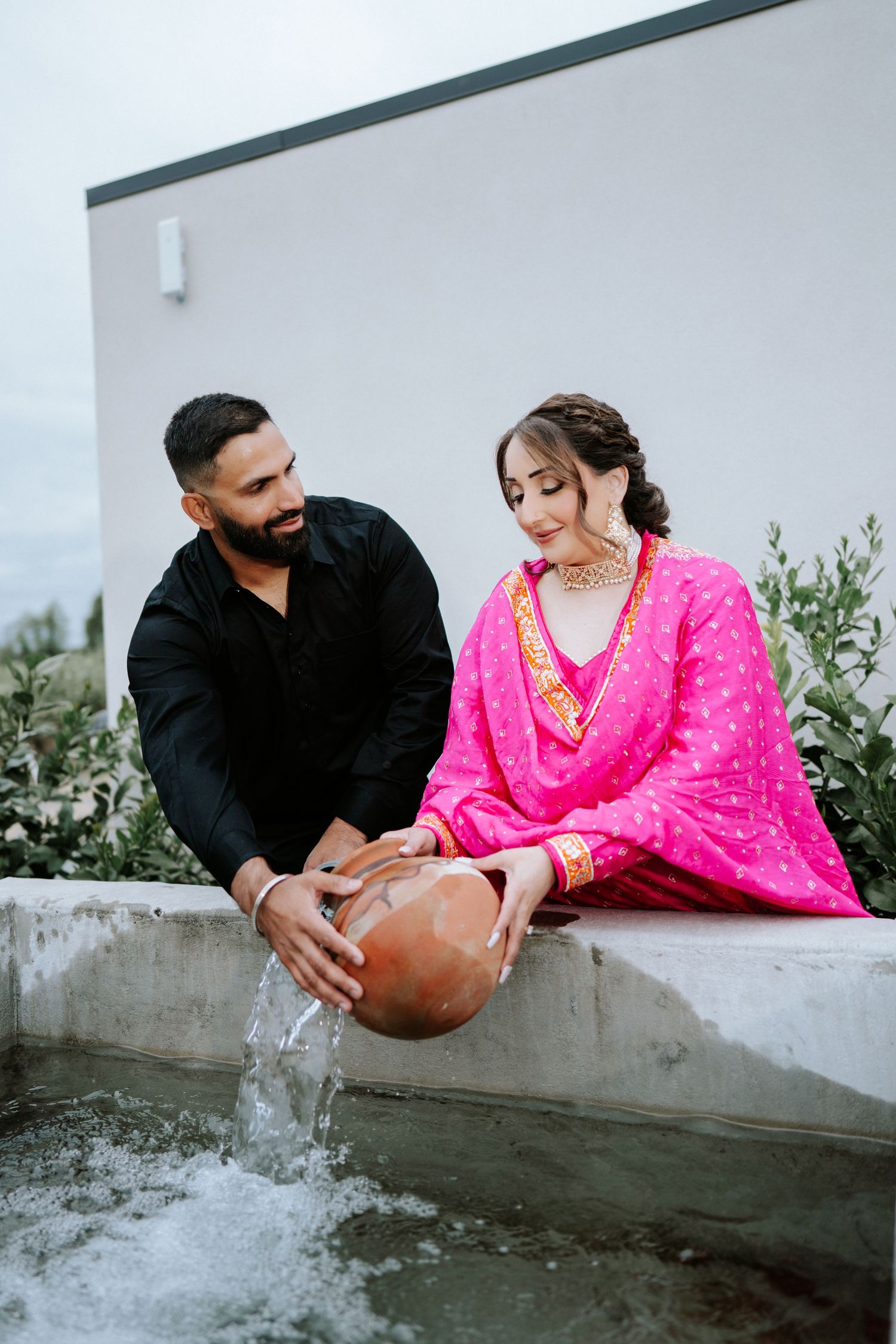 Couple in traditional South Asian attire holding a clay pot near a water feature outdoors.