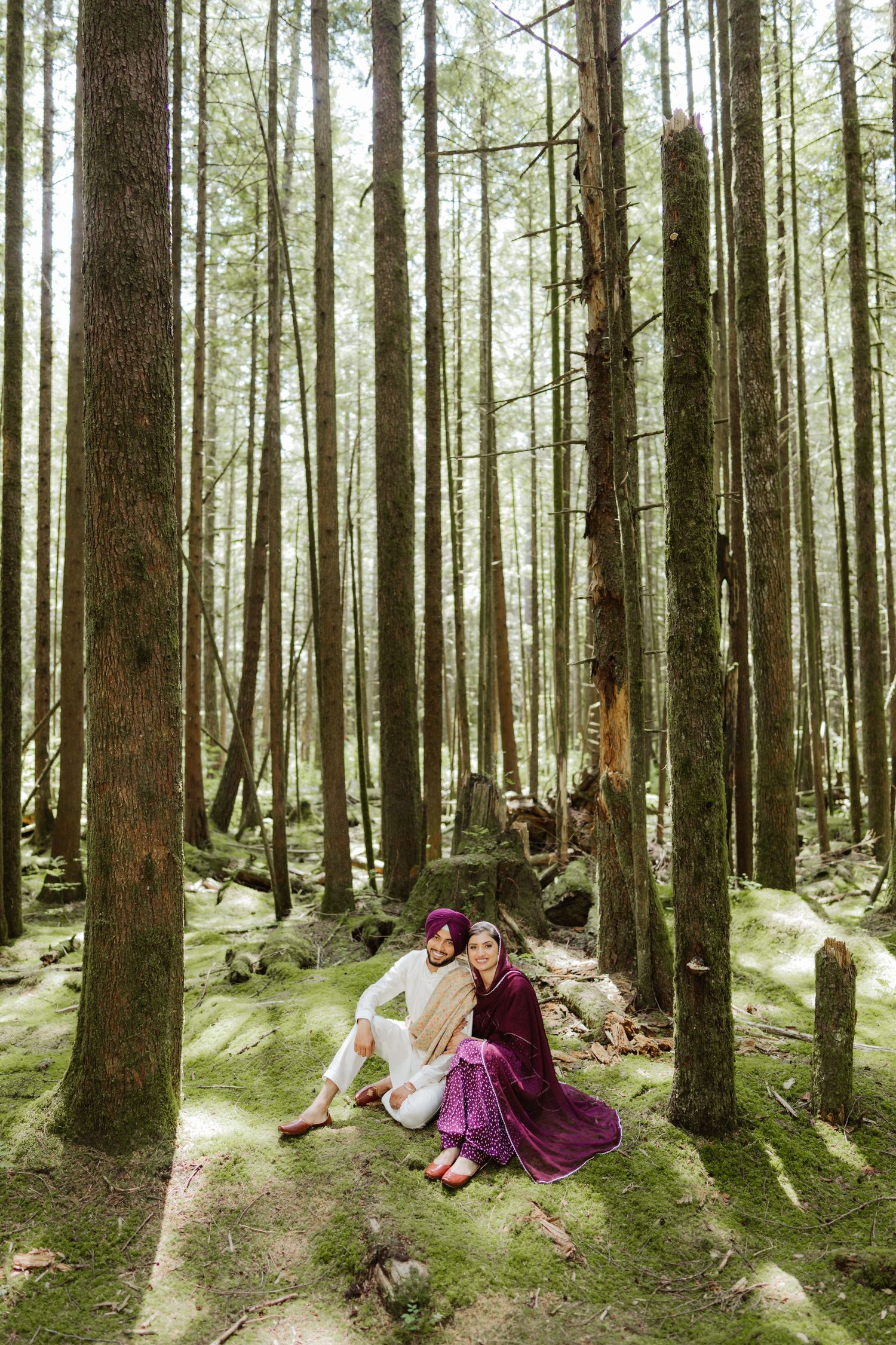 Sikh couple in traditional attire sitting together on mossy ground in a tall forest.