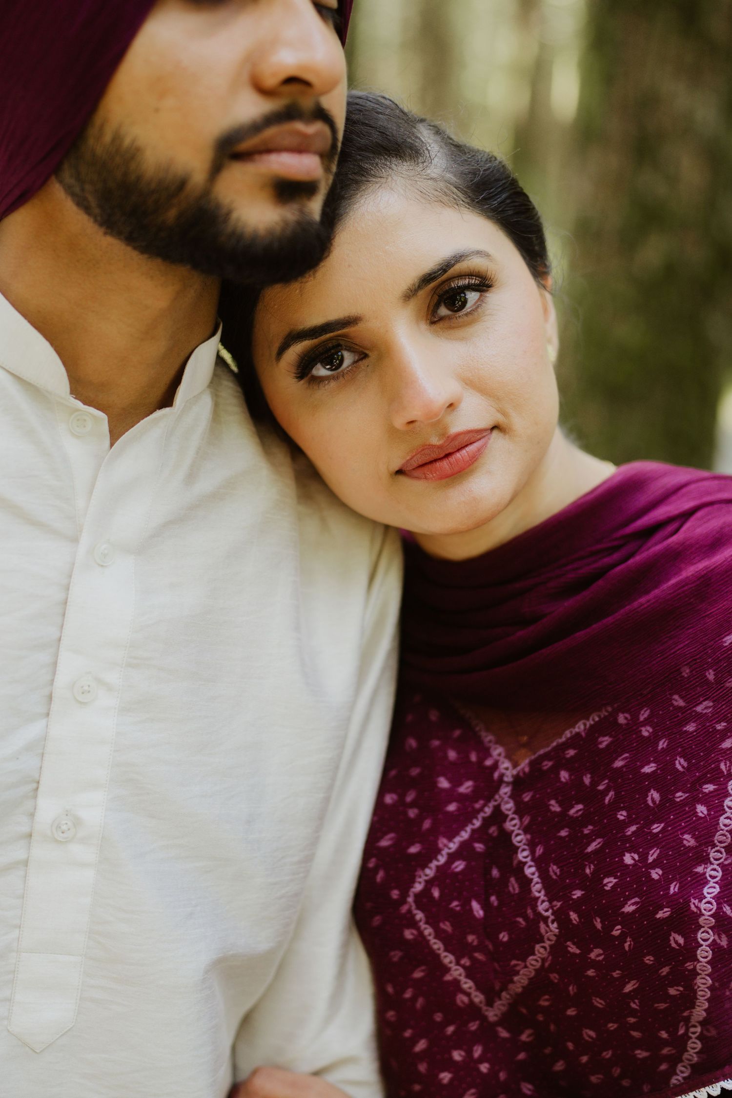 A Sikh couple in traditional attire, woman in burgundy dupatta leaning on man in white kurta and maroon turban.