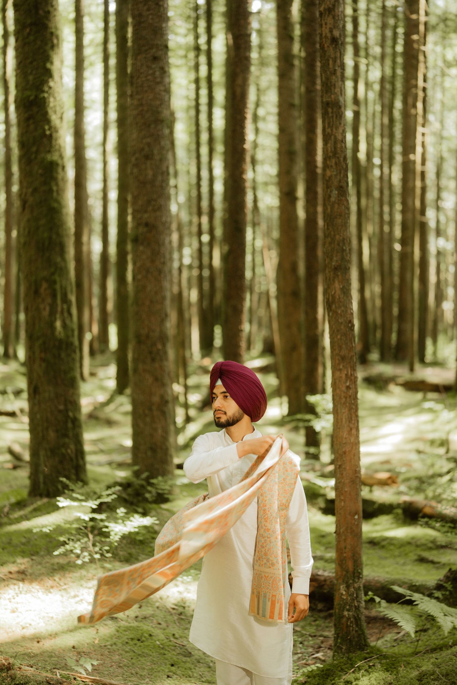 Sikh man in red turban and white kurta holding a golden dupatta scarf while standing in a lush green forest.