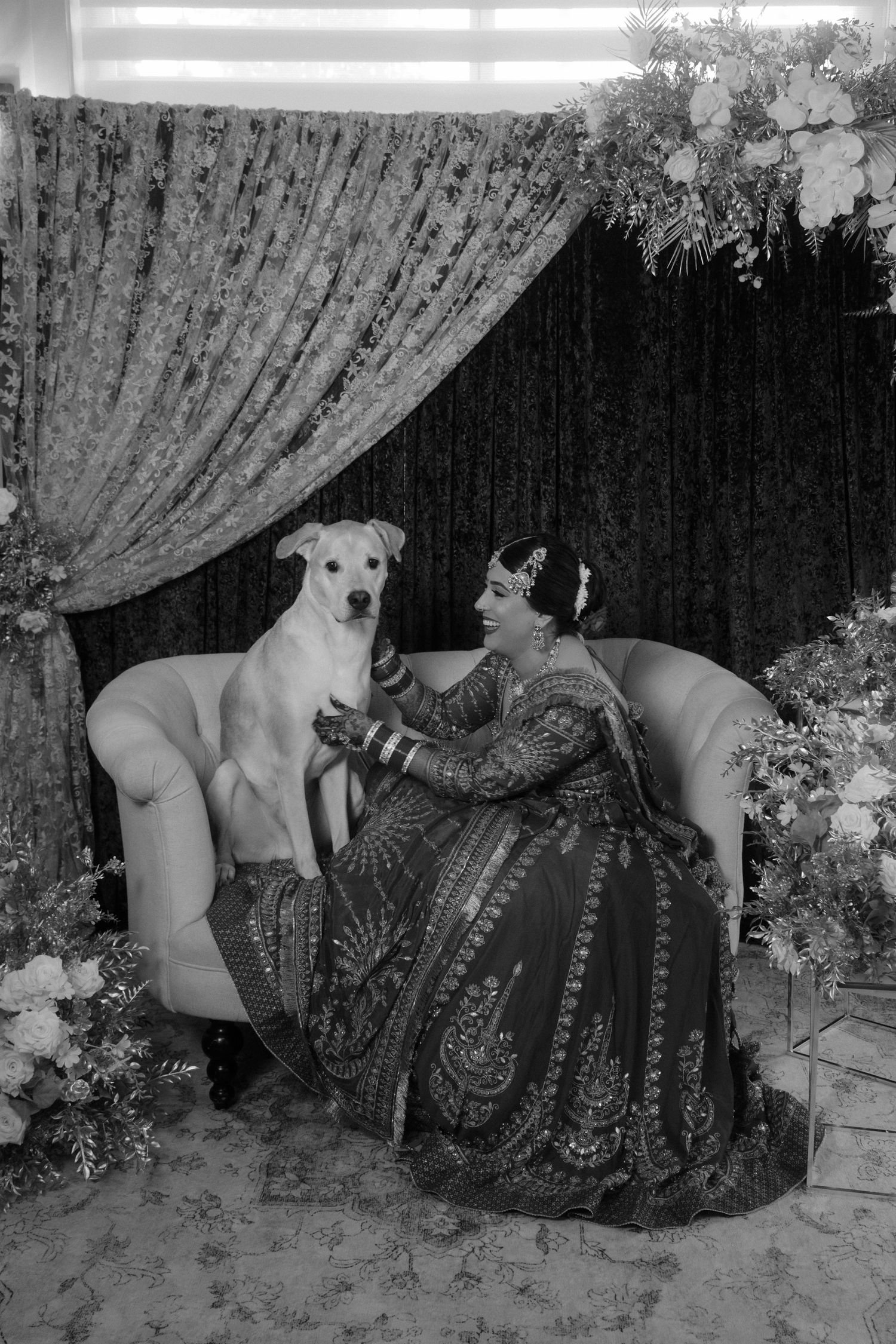 Bride in ornate lehenga sits on sofa with white dog amid floral decor, black and white wedding photo.