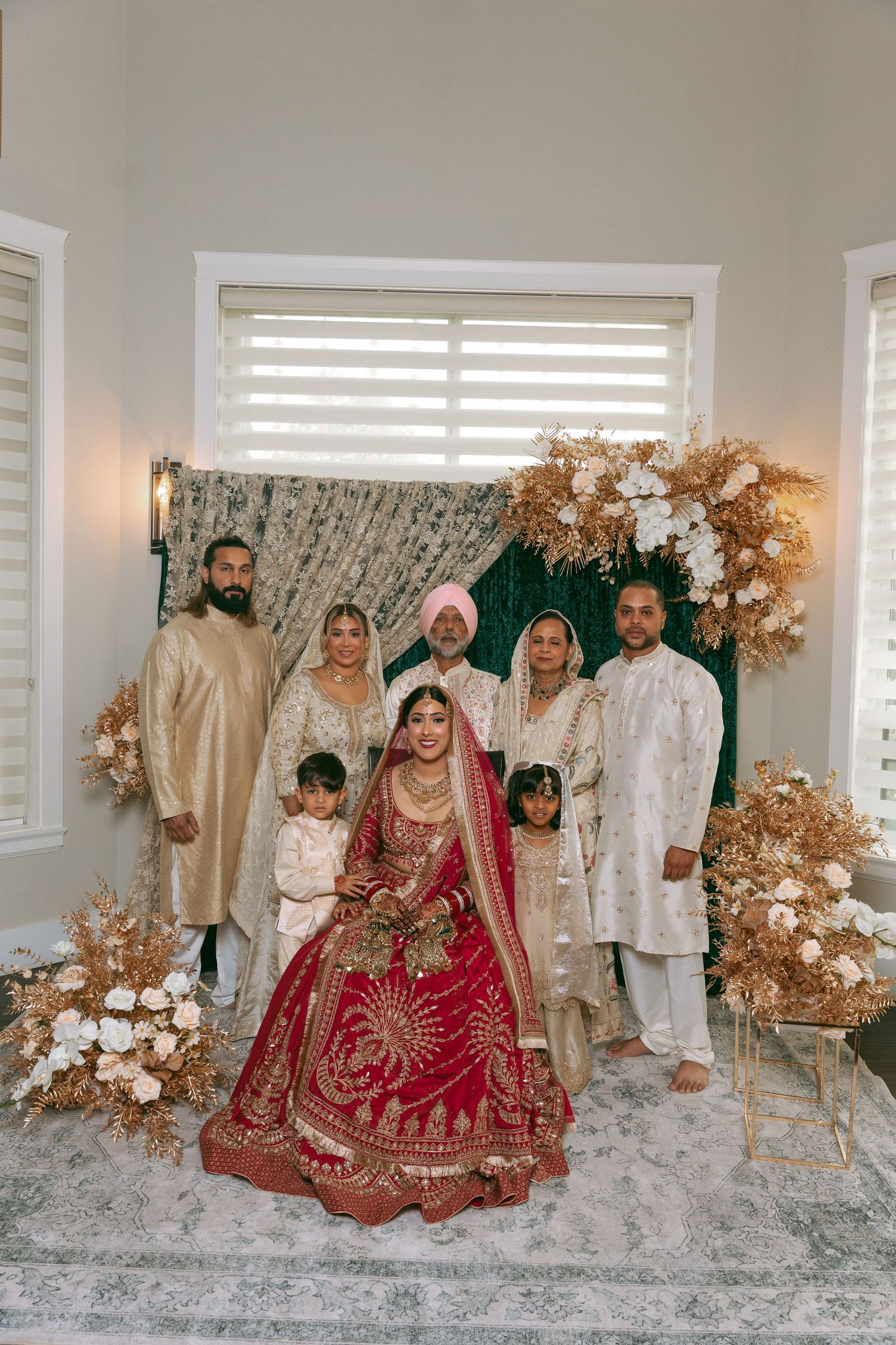 Sikh bride in red lehenga poses with family before floral backdrop at traditional wedding ceremony.
