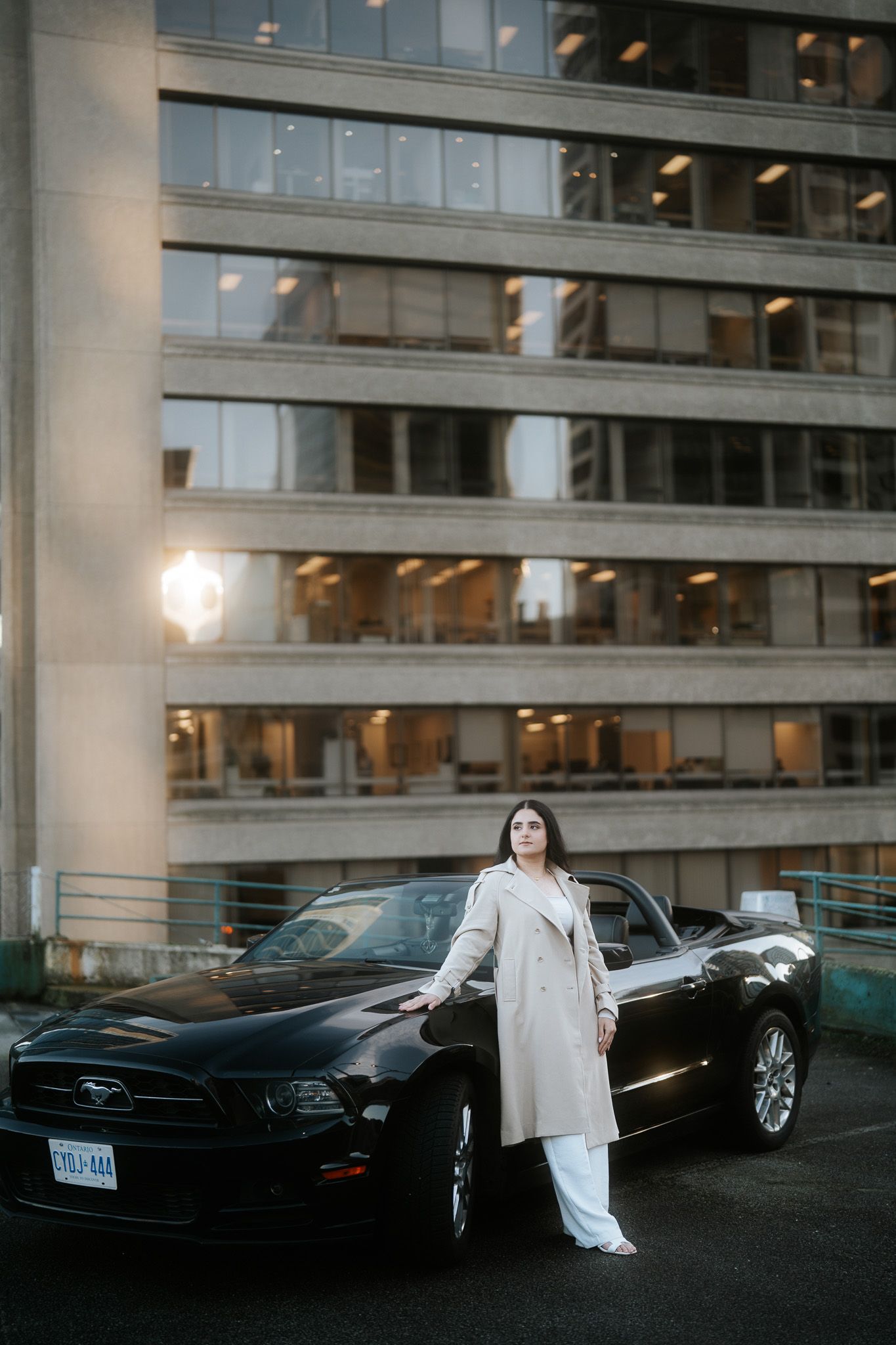 A black Ford Mustang parked outside a modern office building at dusk with someone leaning against it in a cream coat.