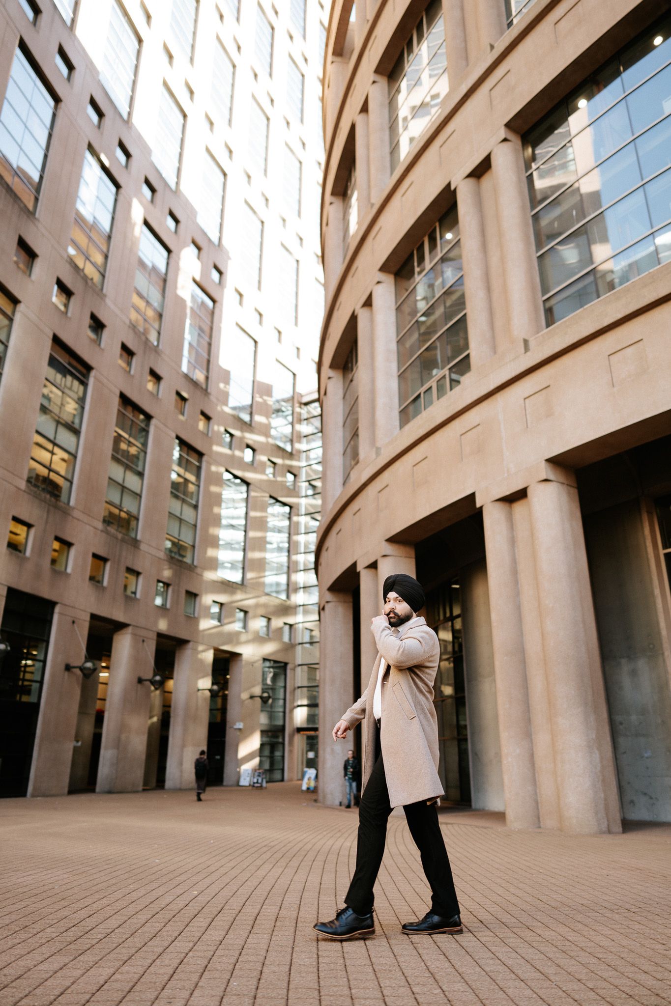 Figure in beige coat stands against curved modern office building with glass windows and stone facade.