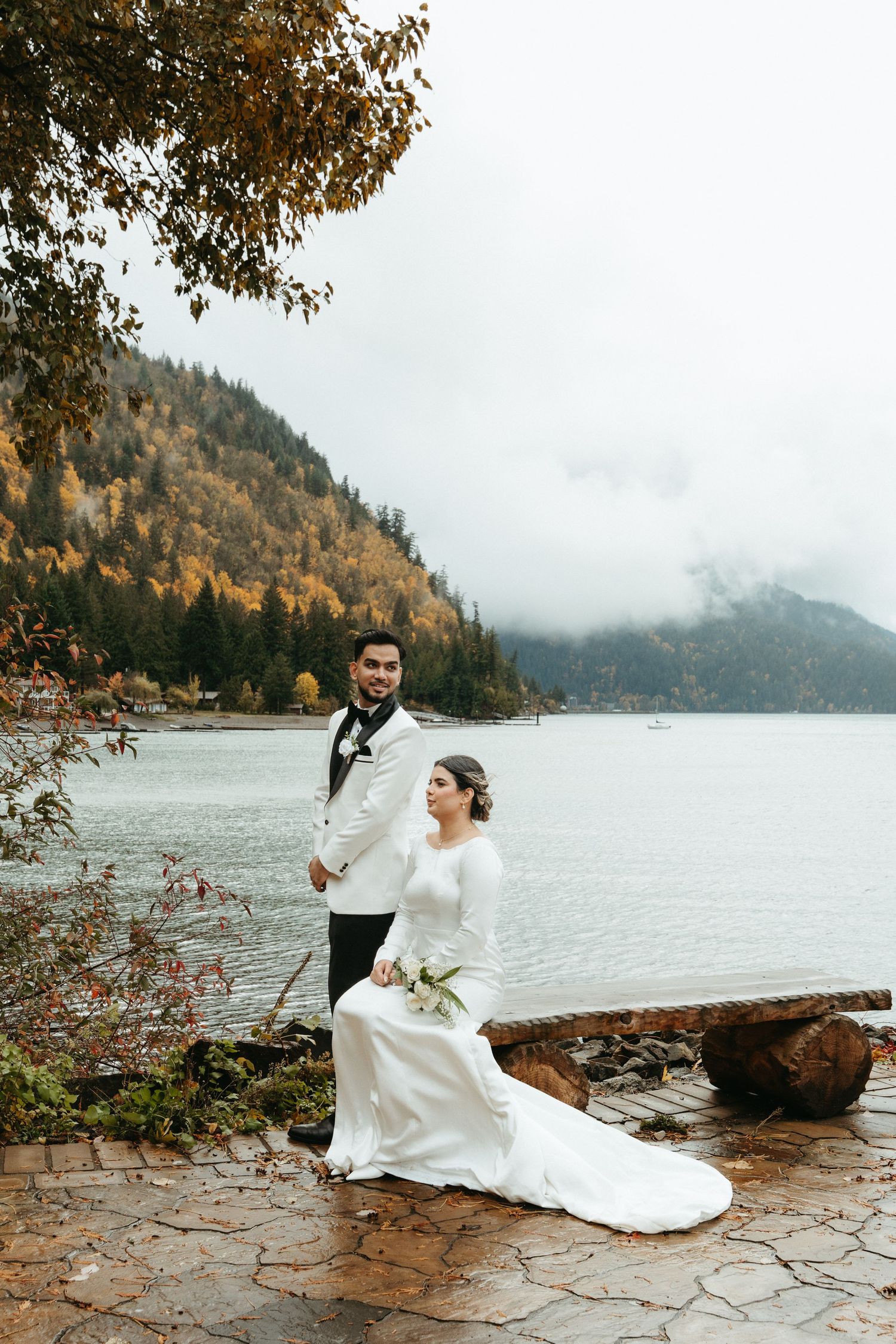 A couple in wedding attire poses along a misty lakeside with autumn foliage and mountains in the background.
