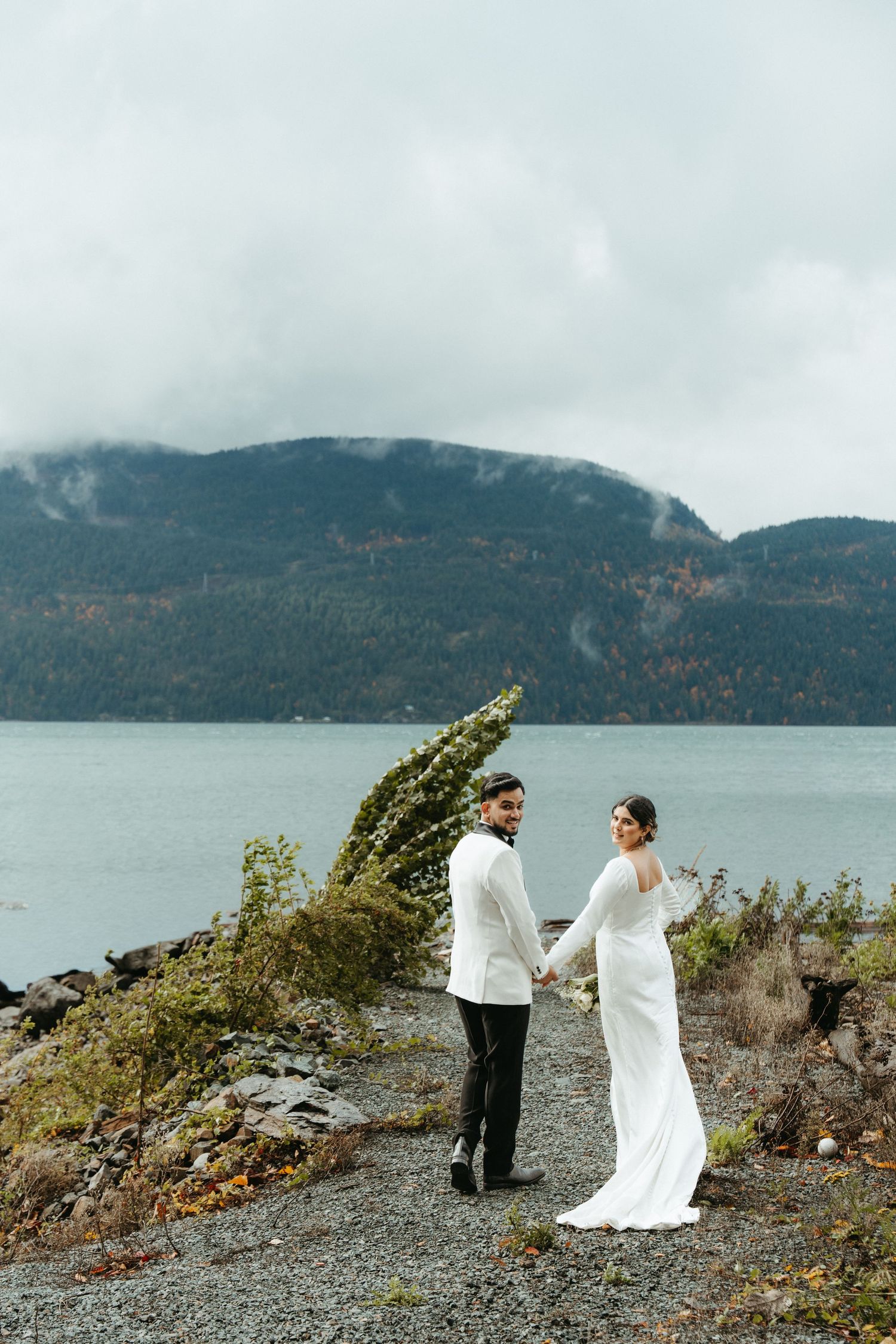 A couple in wedding attire stands on a rocky shoreline with misty mountains and a lake in the background.