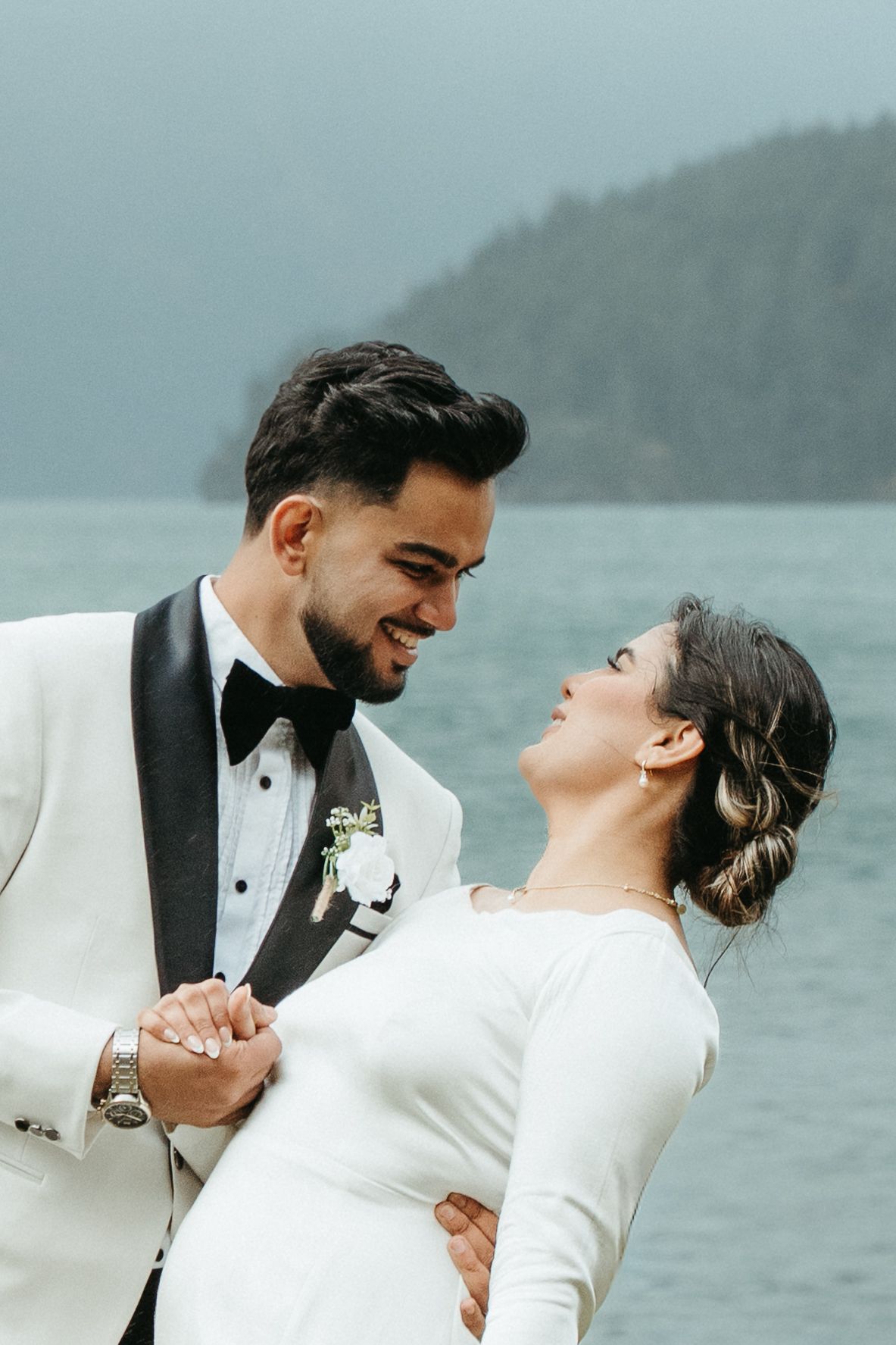 A couple in wedding attire share a romantic moment by a misty lake, with the bride in white and groom in white tuxedo.