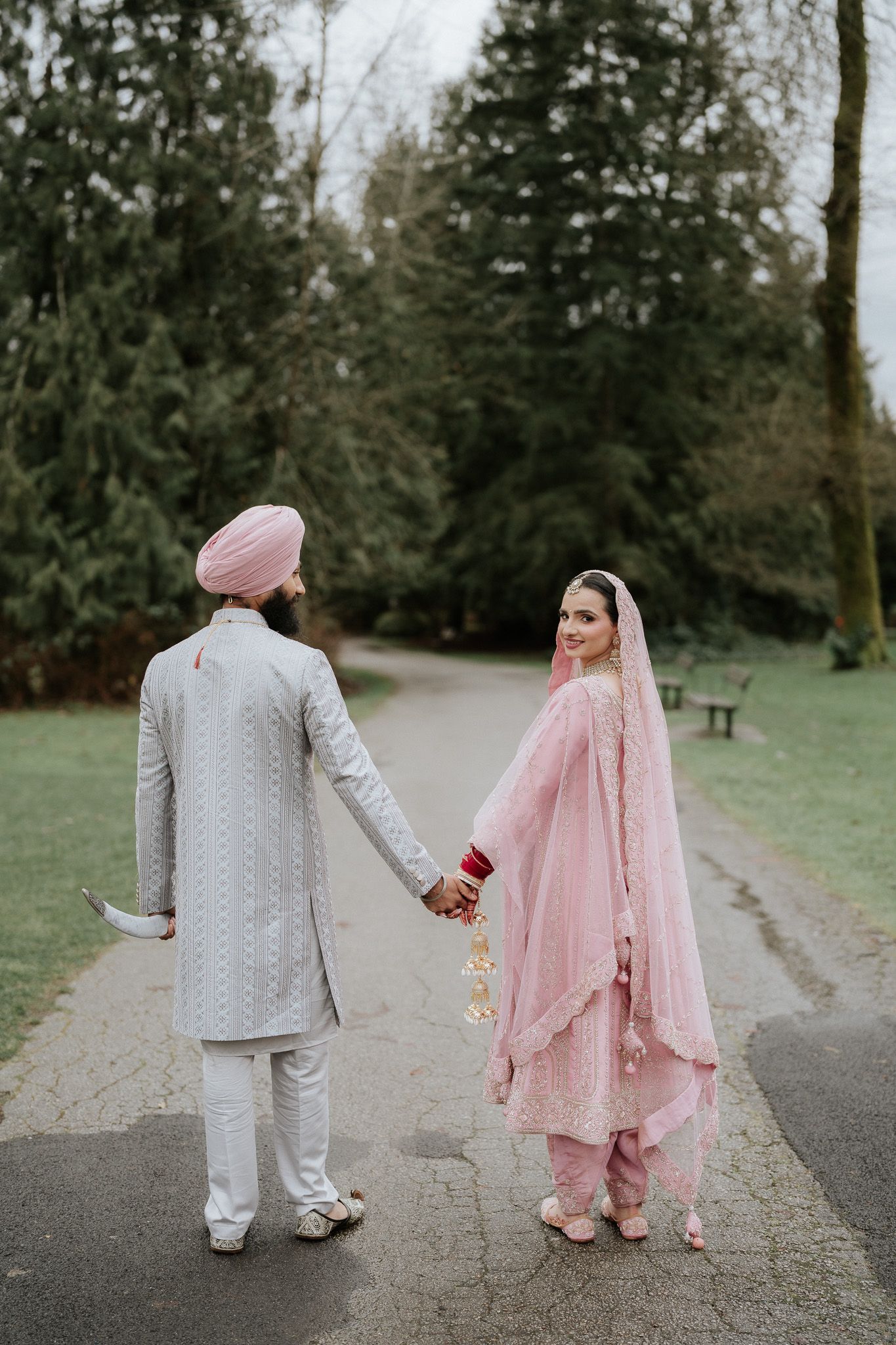 A couple in traditional South Asian wedding attire holding hands on a path surrounded by evergreen trees.