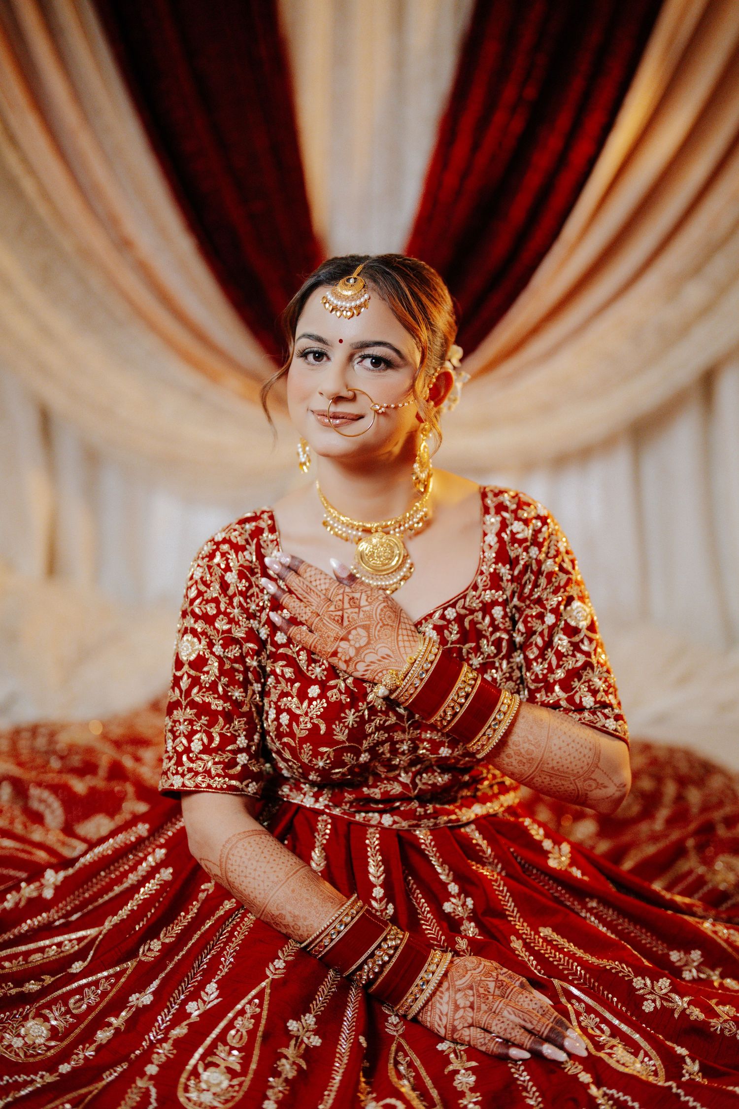 Traditional Indian bridal portrait featuring elaborate red lehenga with gold embellishments against draped backdrop.