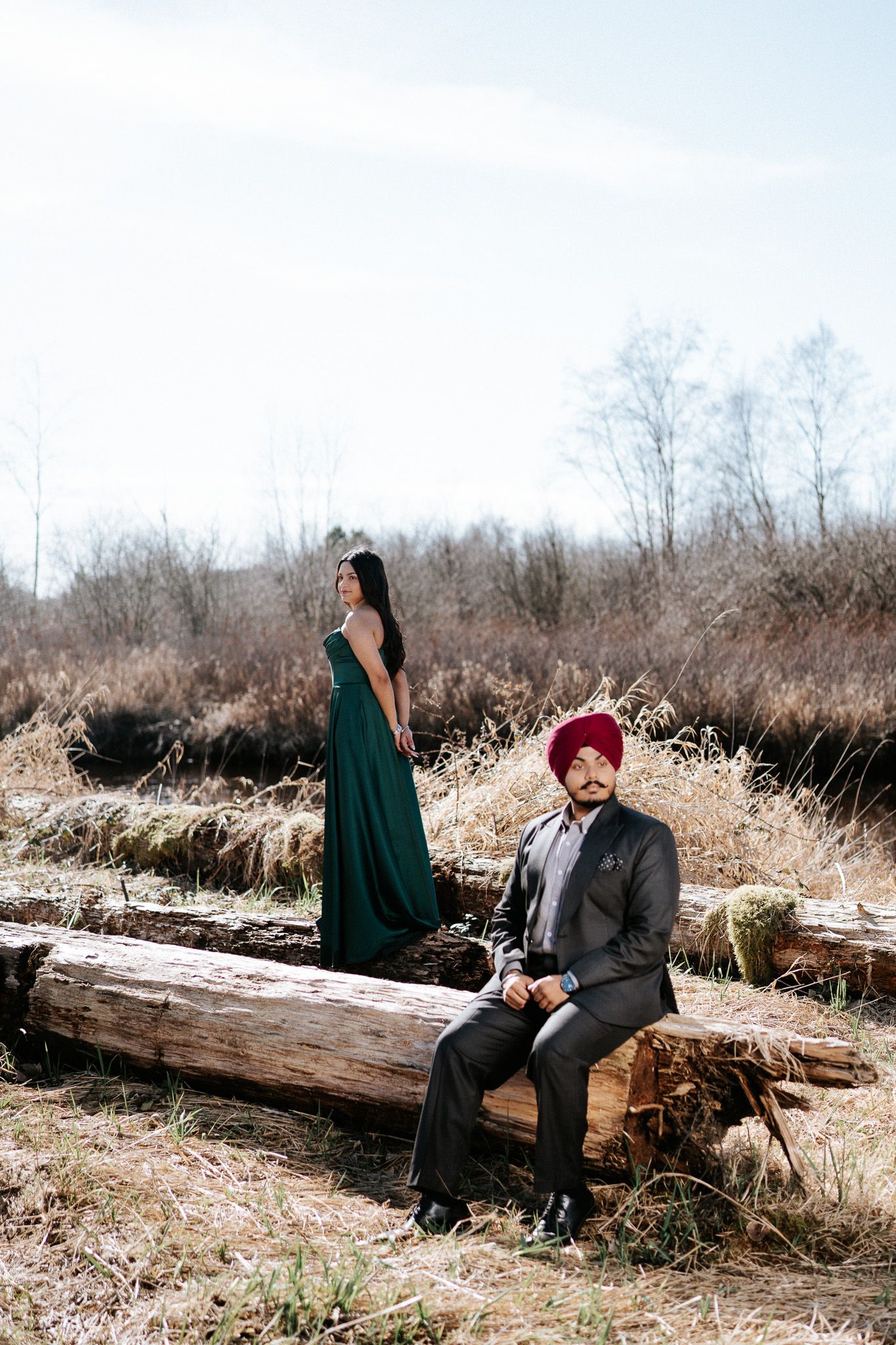 A couple sits together on a rustic wooden log bench in a winter field with dried grass and bare trees.