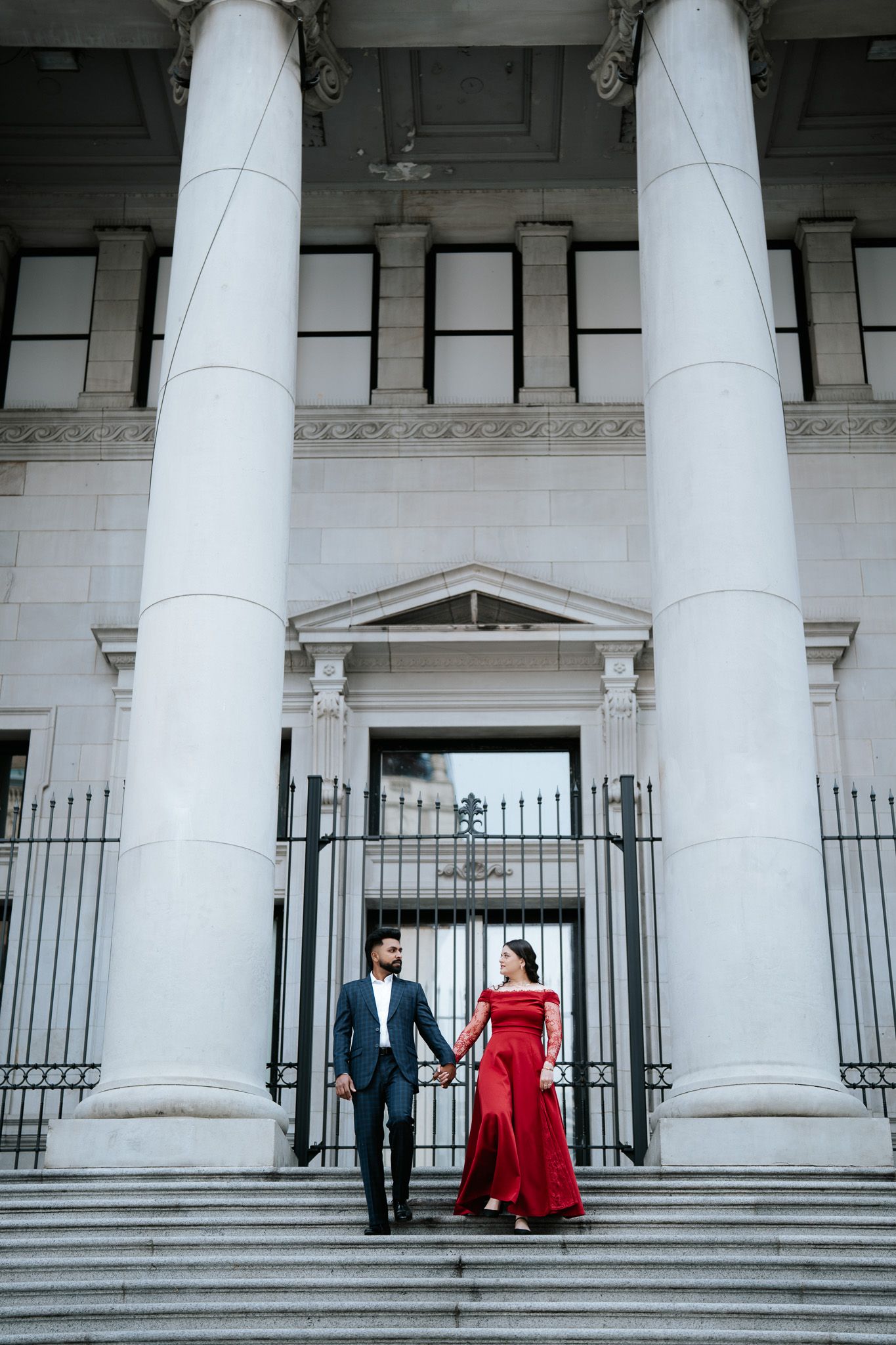 Couple holding hands on grand building steps, man in navy suit, woman in red off-shoulder gown.