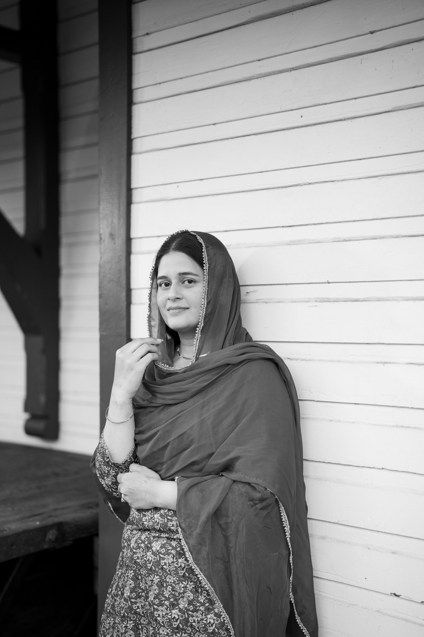 Black and white portrait of a young South Asian woman in a traditional dupatta leaning against a white wooden wall.
