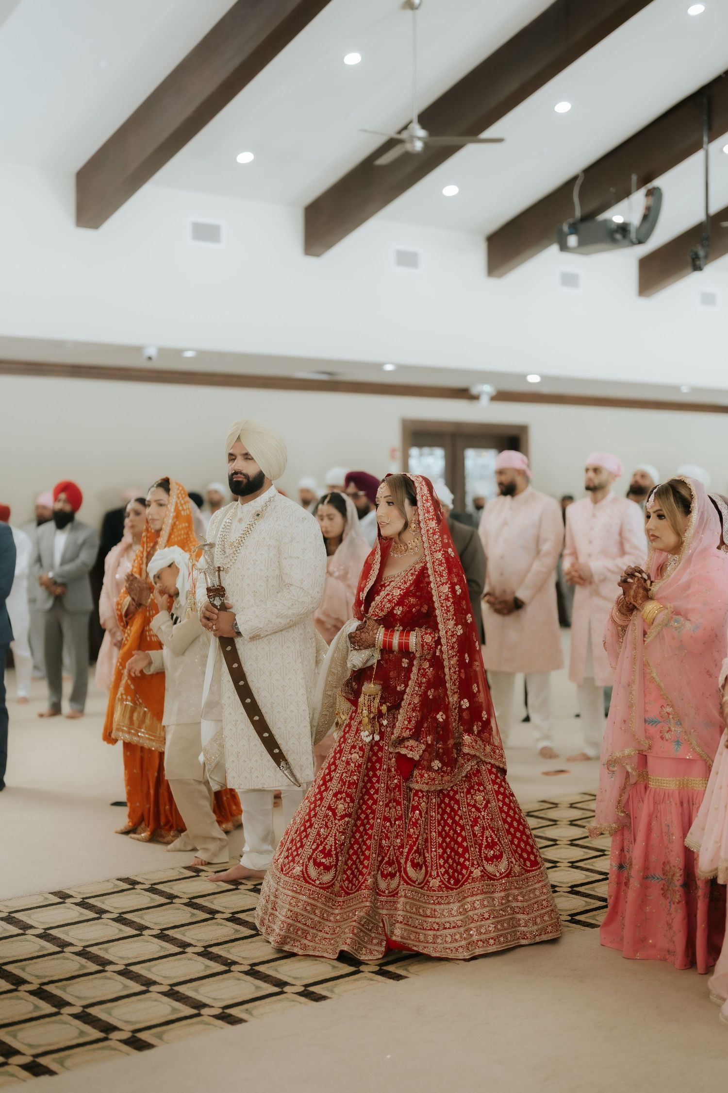 Sikh wedding ceremony with bride in red lehenga and groom in white sherwani processing through a decorated hall.
