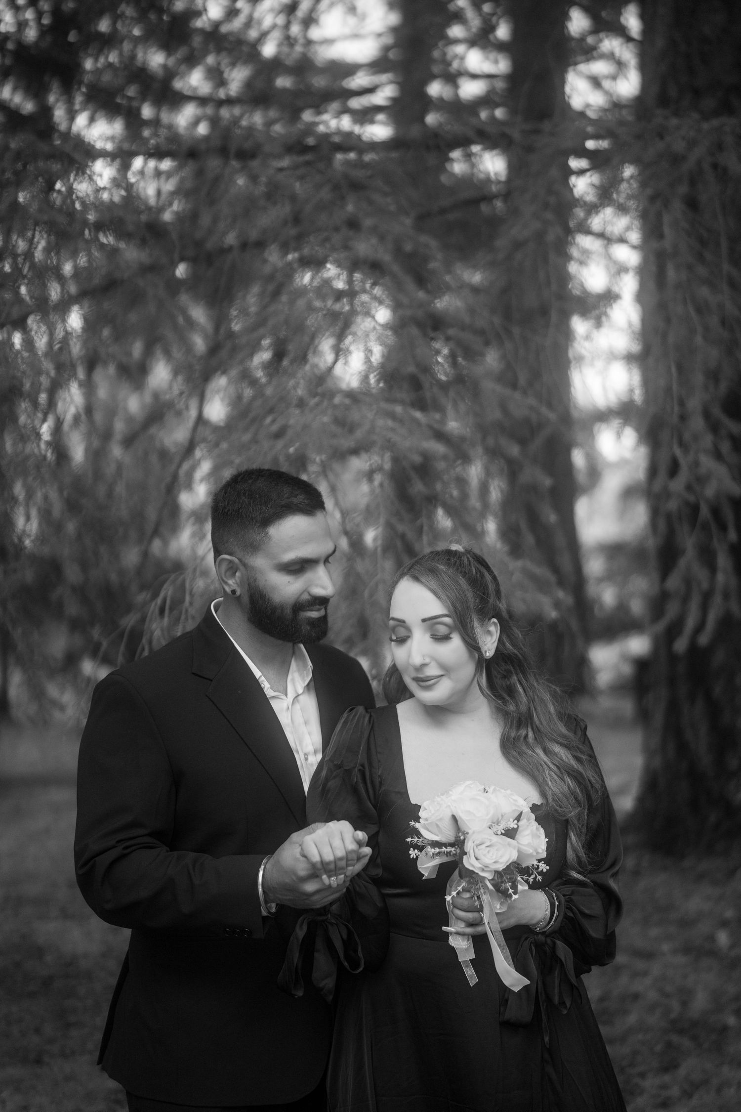 Black and white photo of a bearded groom in suit and bride holding bouquet standing together in a forest setting.