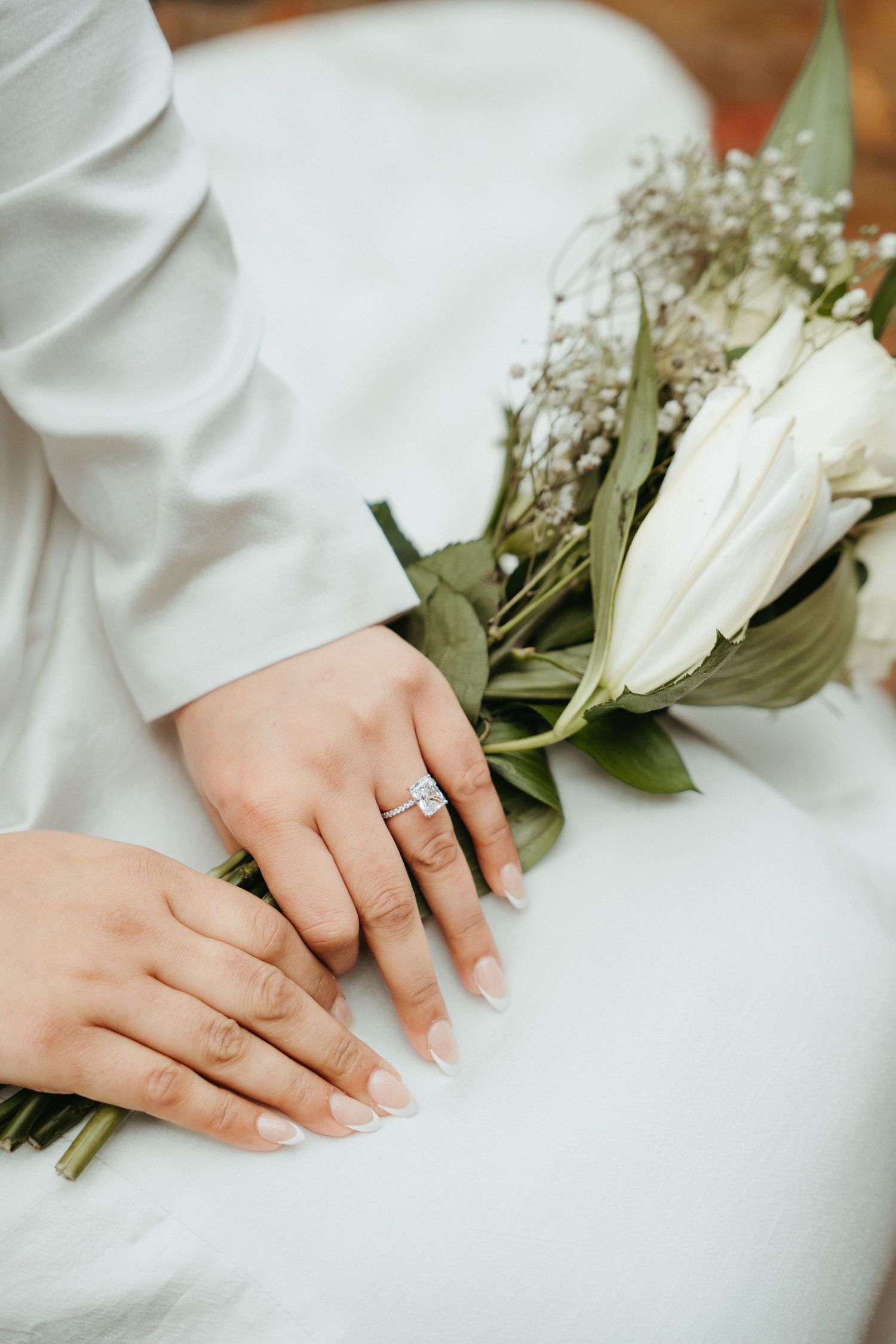Hand wearing an elegant engagement ring holds white roses against a soft white fabric background.