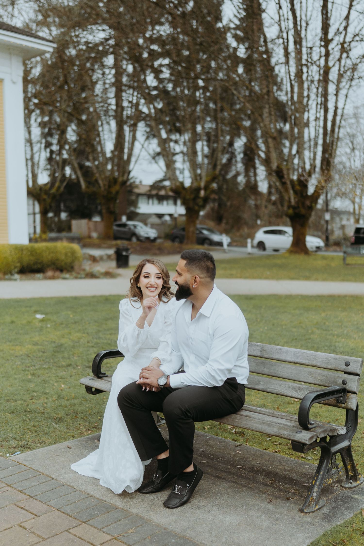 A couple dressed in white and black sits close together on a wooden bench in front of a church.
