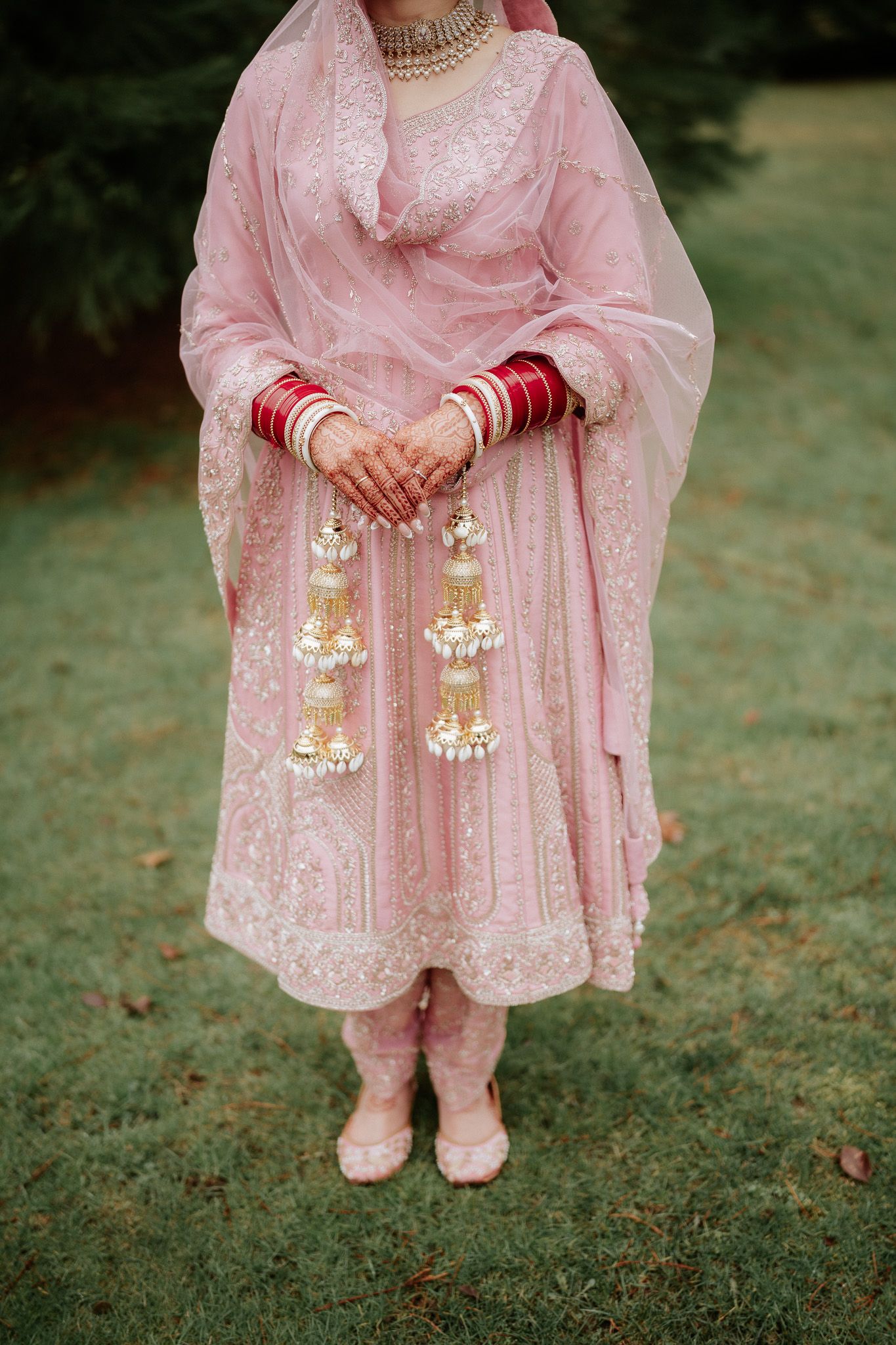 Close-up details of pink traditional Indian bridal attire with gold embroidery and matching accessories on grass.