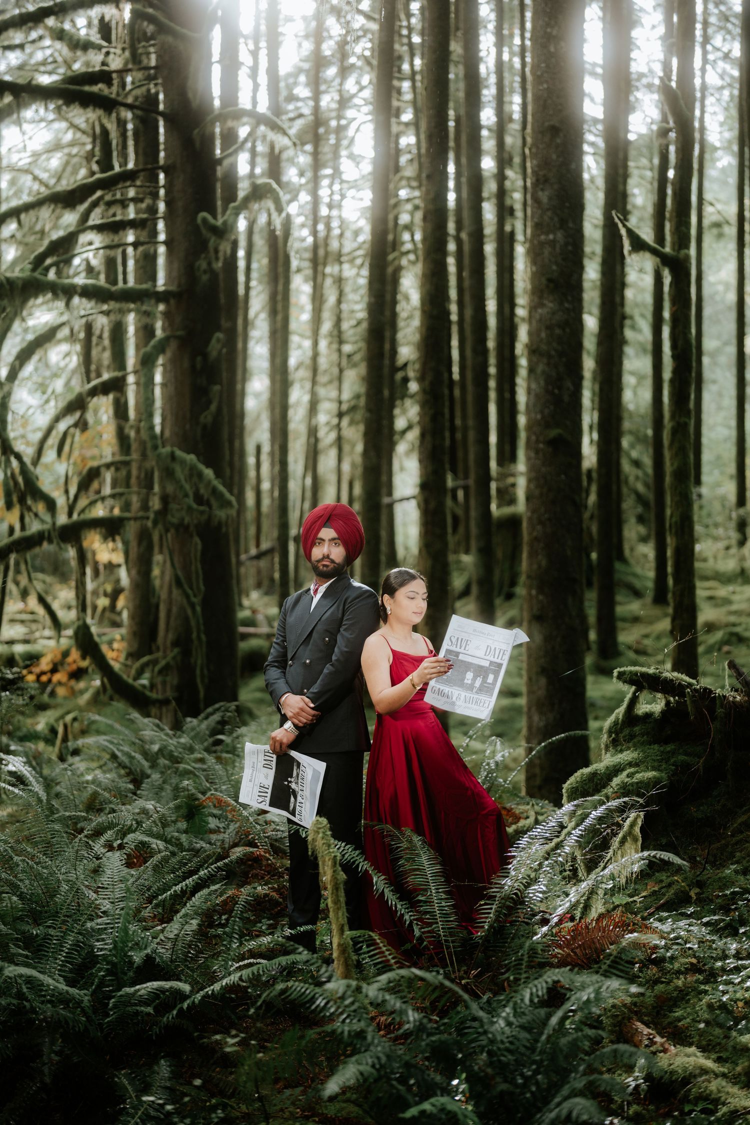 A couple in red and black formal wear poses with newspapers among tall trees and ferns.