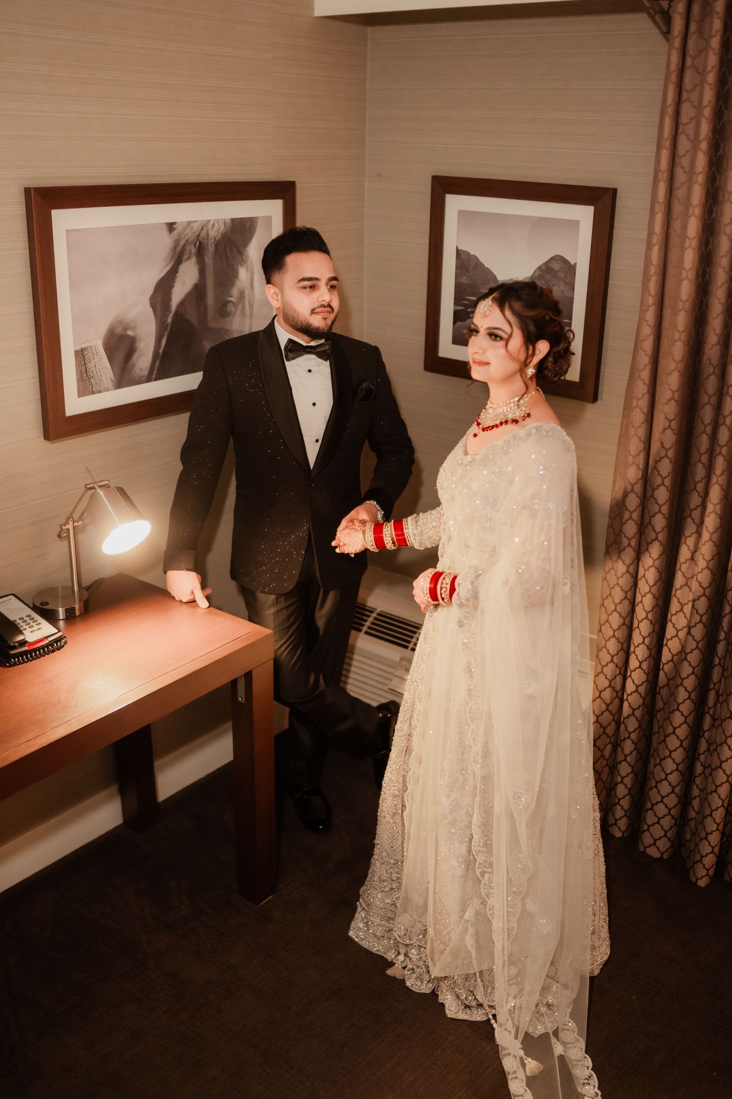 A couple in formal attire shares moments in a hotel room with brown curtains.