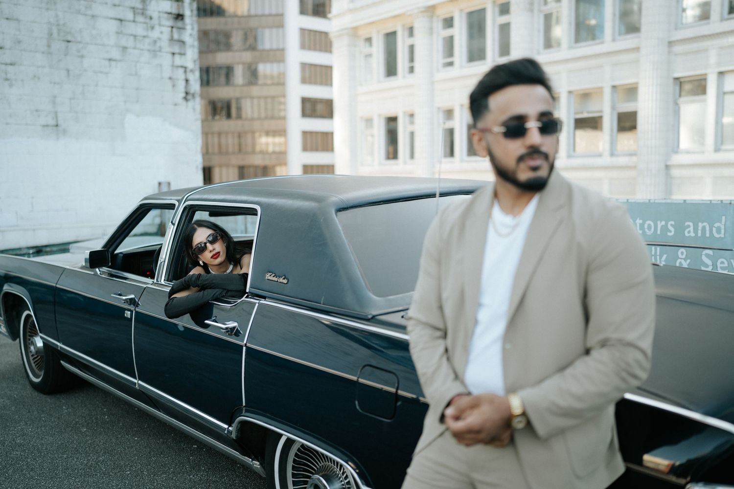 A vintage black convertible car parked on a city street with someone in a beige suit and sunglasses nearby.