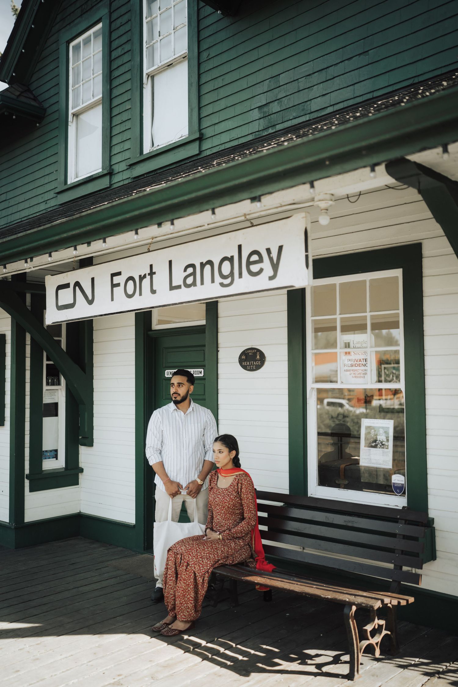 A couple sits on a wooden bench outside a historic green and white train station building at Fort Langley.