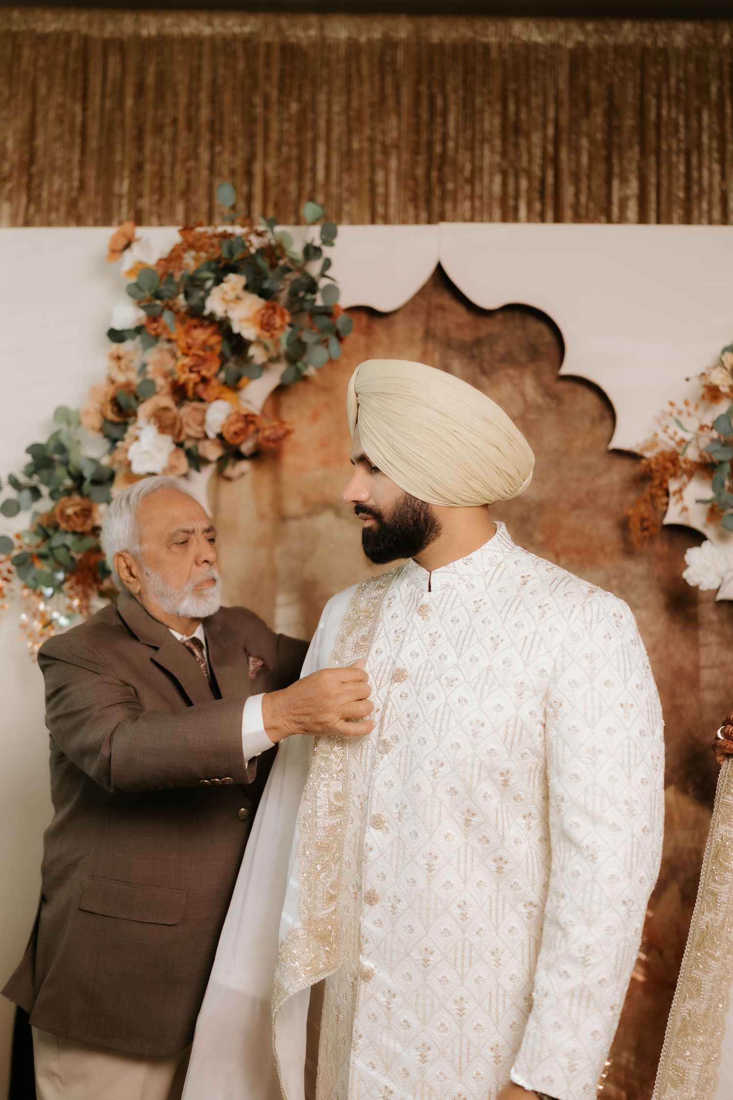 An elderly man adjusts the sherwani of a Sikh groom wearing a cream turban at a traditional wedding ceremony.