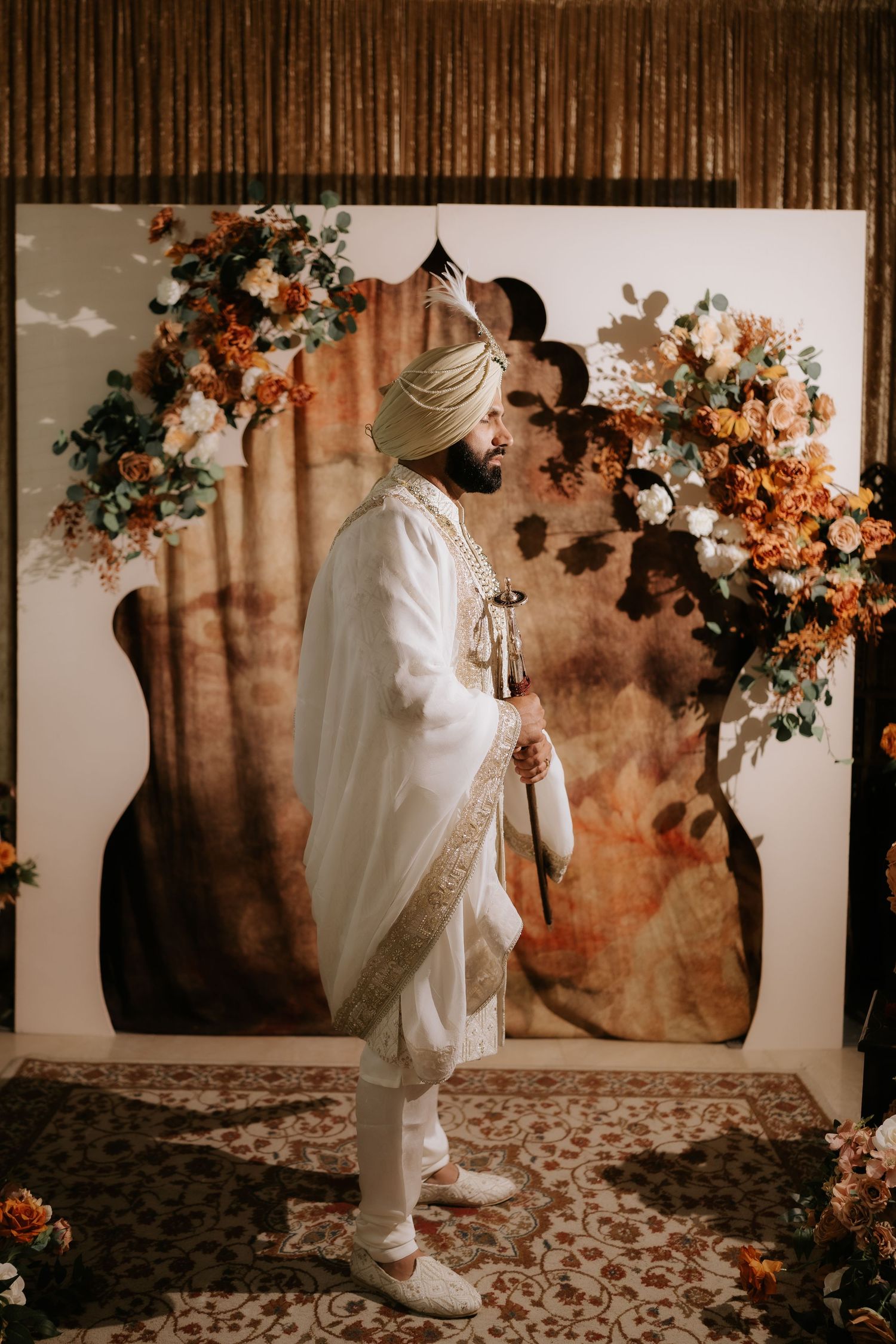 Sikh groom in white sherwani and turban stands before floral backdrop with orange blooms at elegant wedding ceremony.