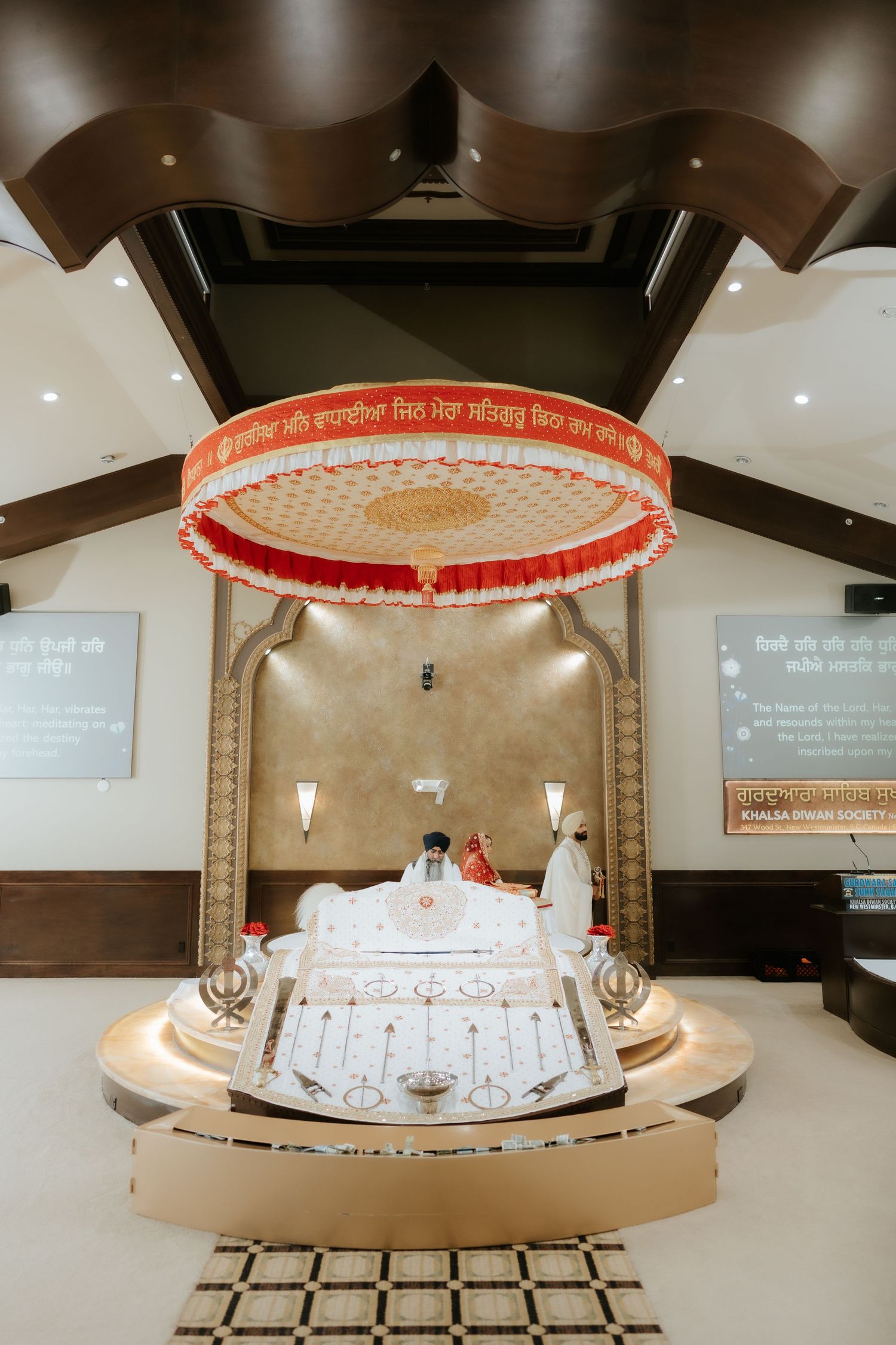 Sikh Gurdwara interior with ornate Palki Sahib throne, red canopy, and Guru Granth Sahib on elevated platform.