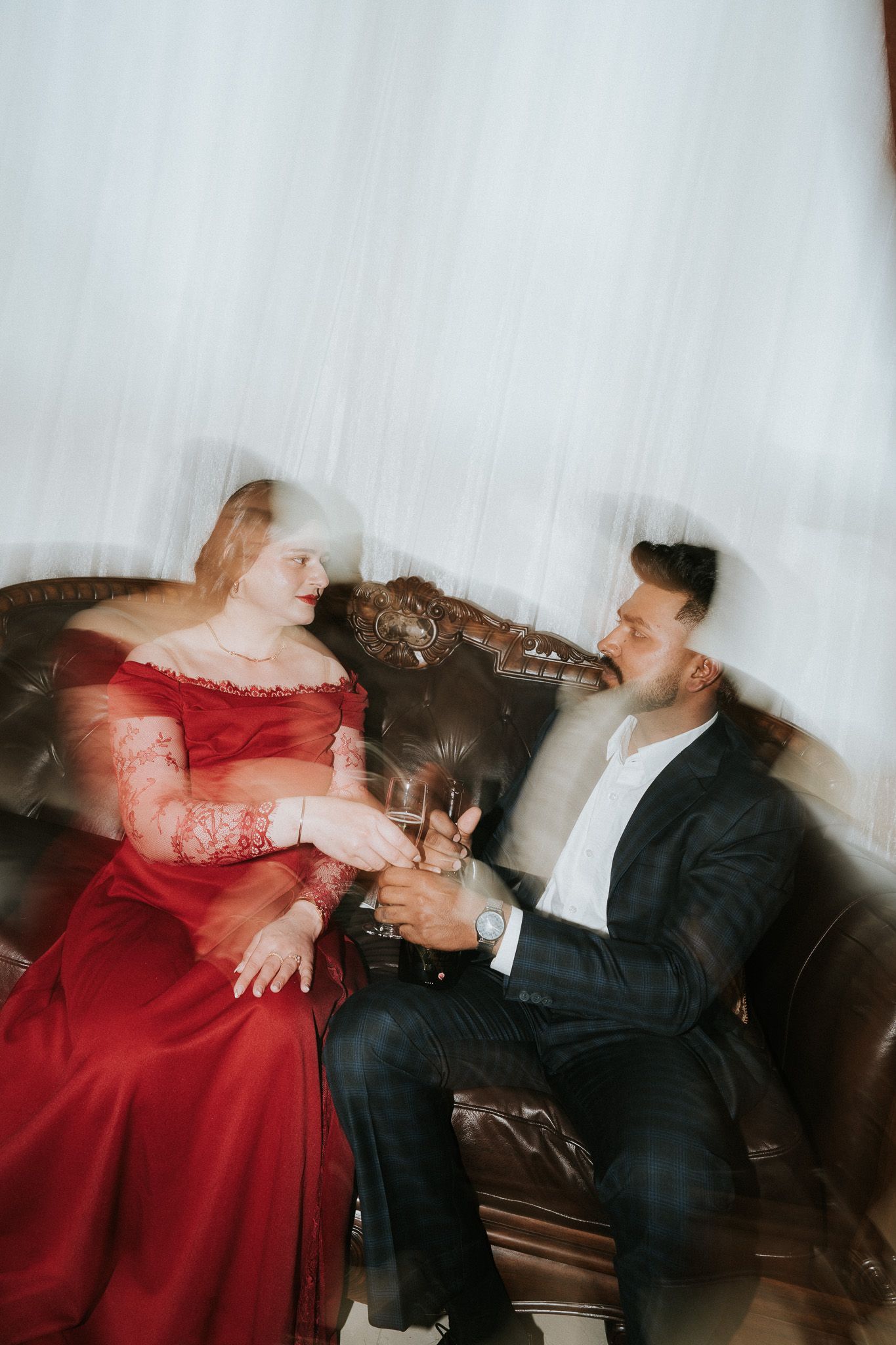 Couple laughing together on leather sofa with champagne glasses, woman in red gown, man in dark suit, artistic blur effect.