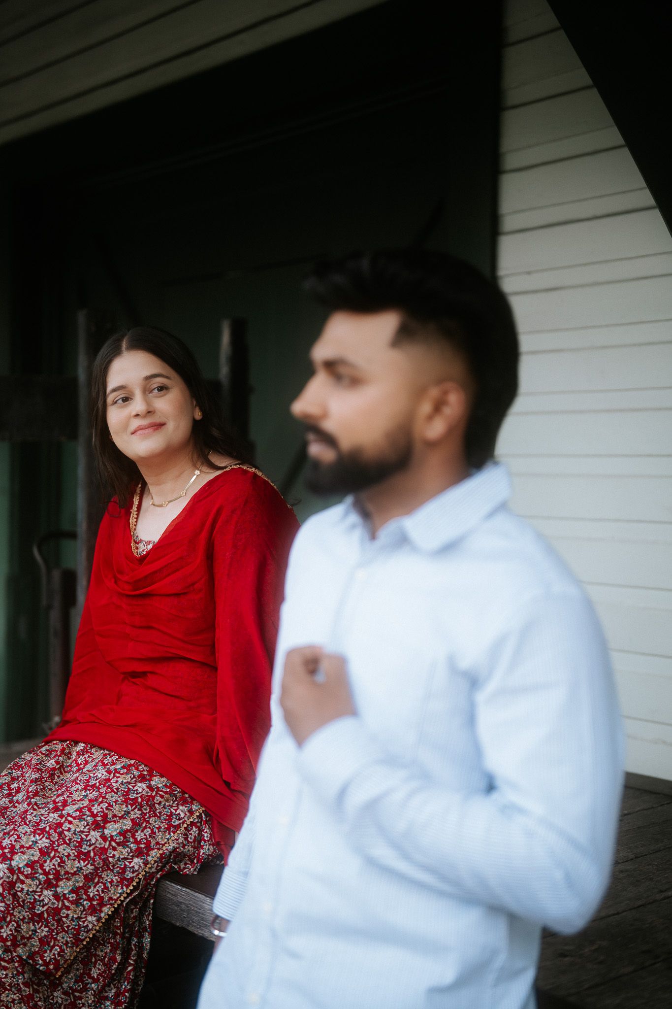 Woman in red salwar kameez sits smiling while bearded man in white shirt stands in foreground outdoors.
