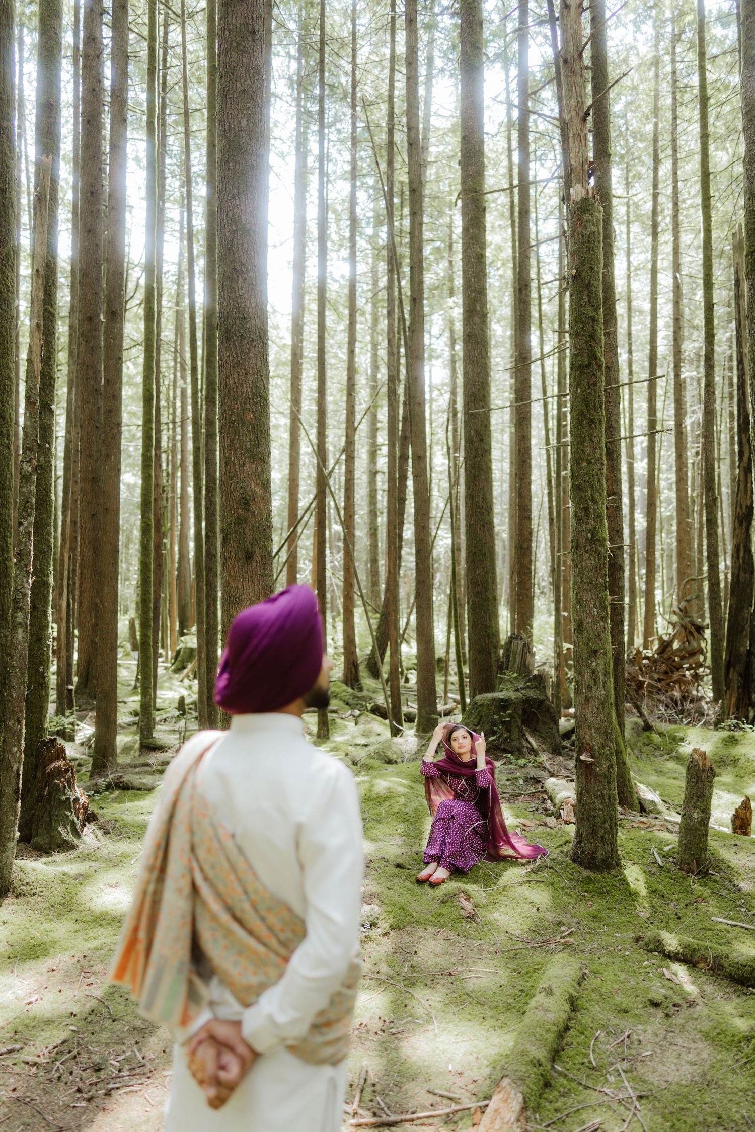 Sikh groom in white kurta approaches bride sitting among tall trees in a sunlit forest.
