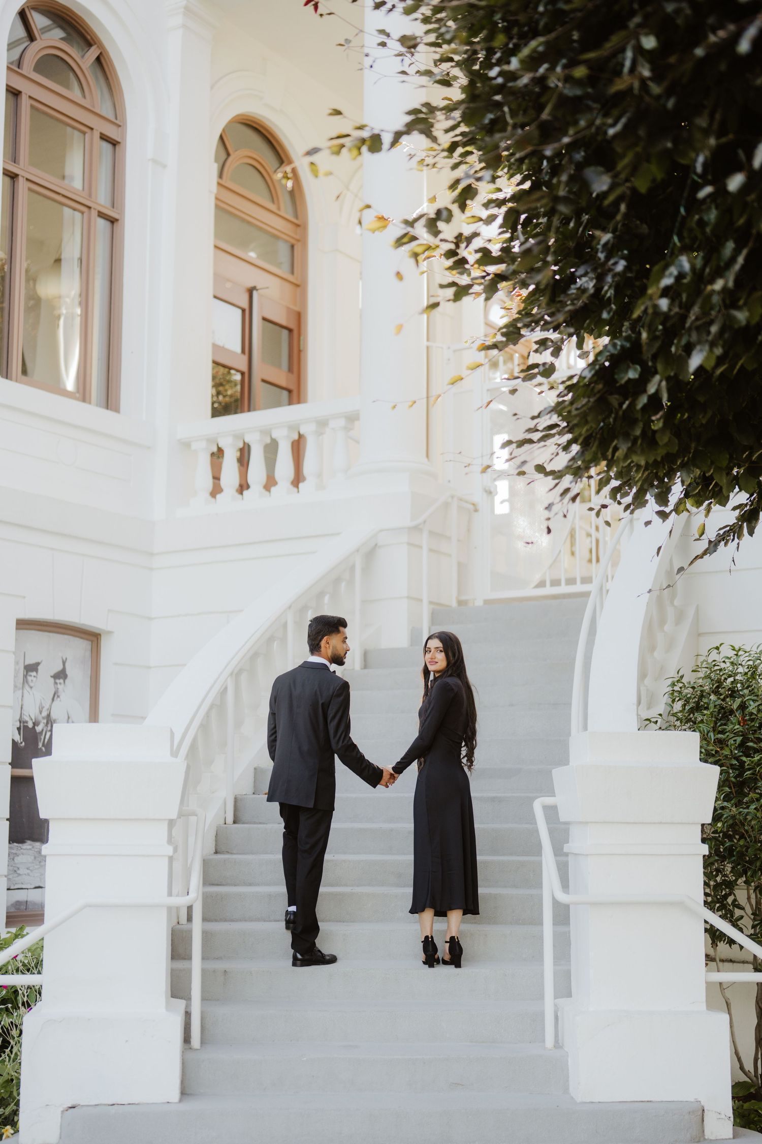 Couple in black formal wear holding hands on white marble staircase outside elegant white building.