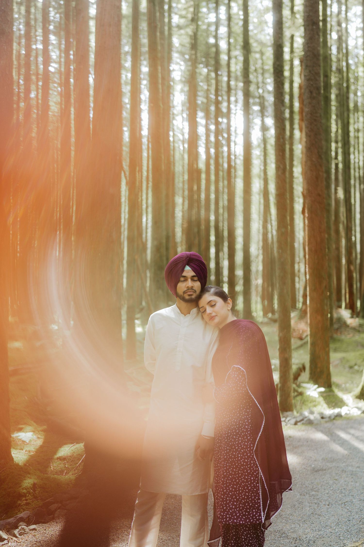 Sikh couple embracing in sunlit forest, man in white kurta and burgundy turban, woman in dark floral dress.