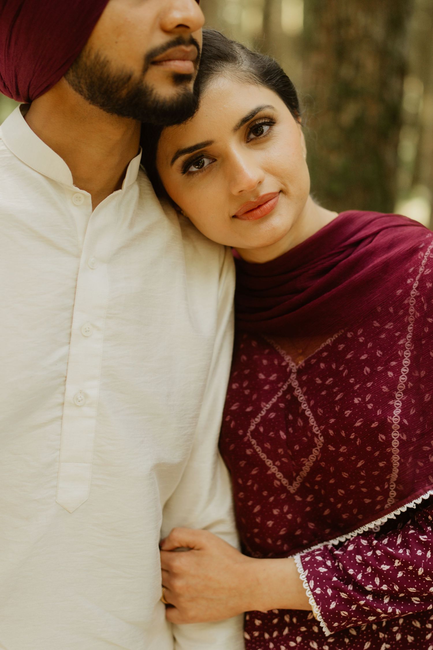 A Sikh couple in traditional attire, woman in maroon salwar kameez leans on man in white kurta and red turban.
