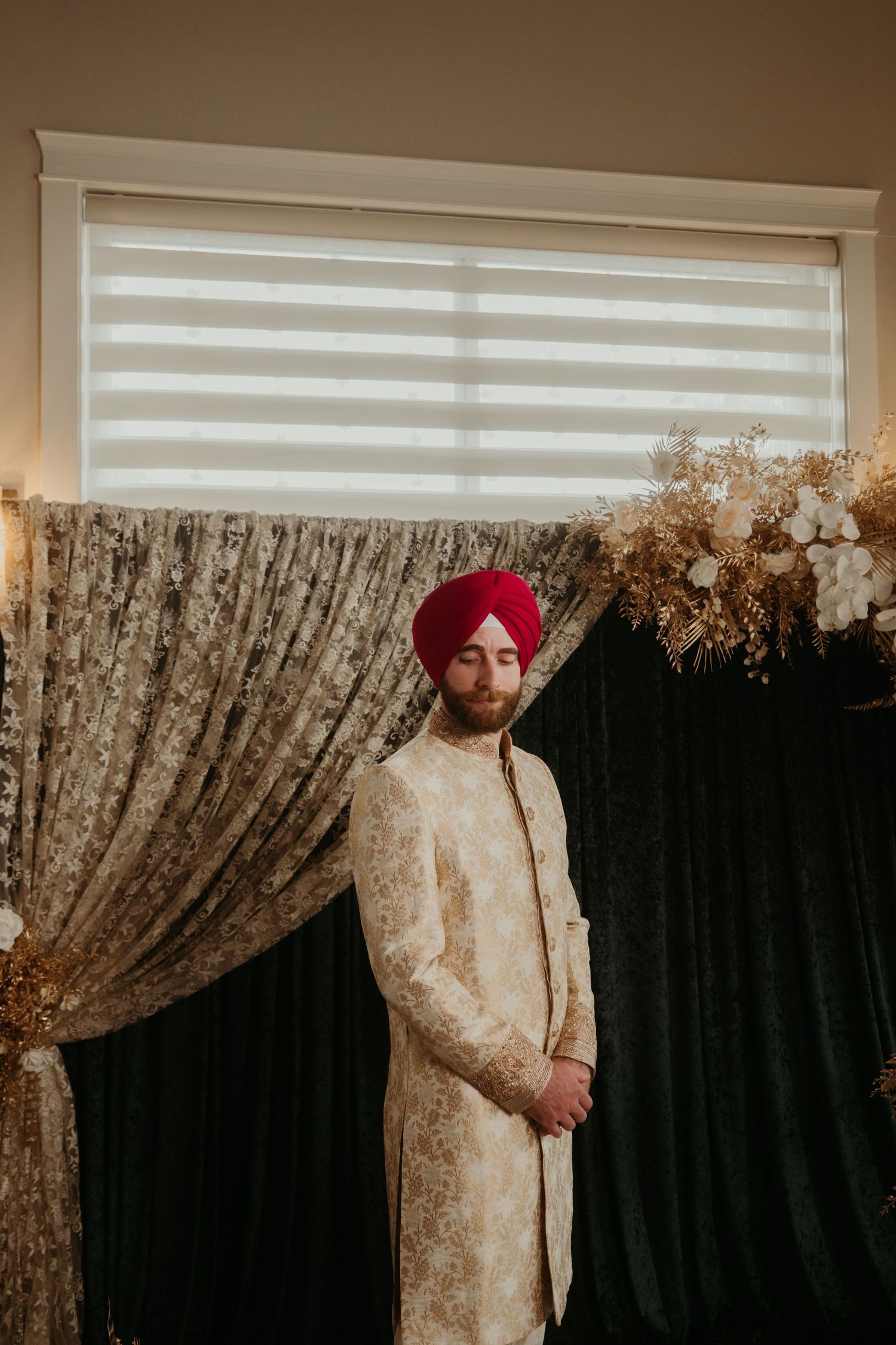 Sikh groom in a red turban and ivory sherwani stands elegantly before draped curtains and floral decor.