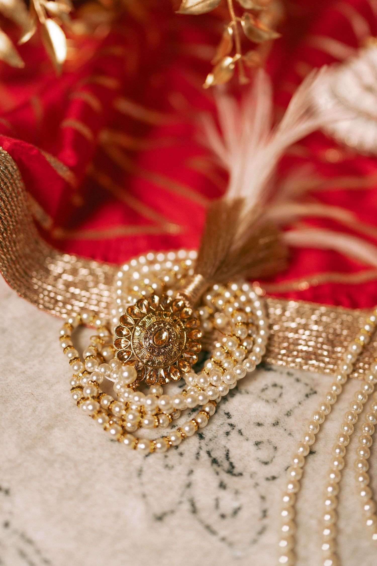 Ornate gold brooch with pearl strands on ivory embroidered fabric, red silk and feather backdrop.