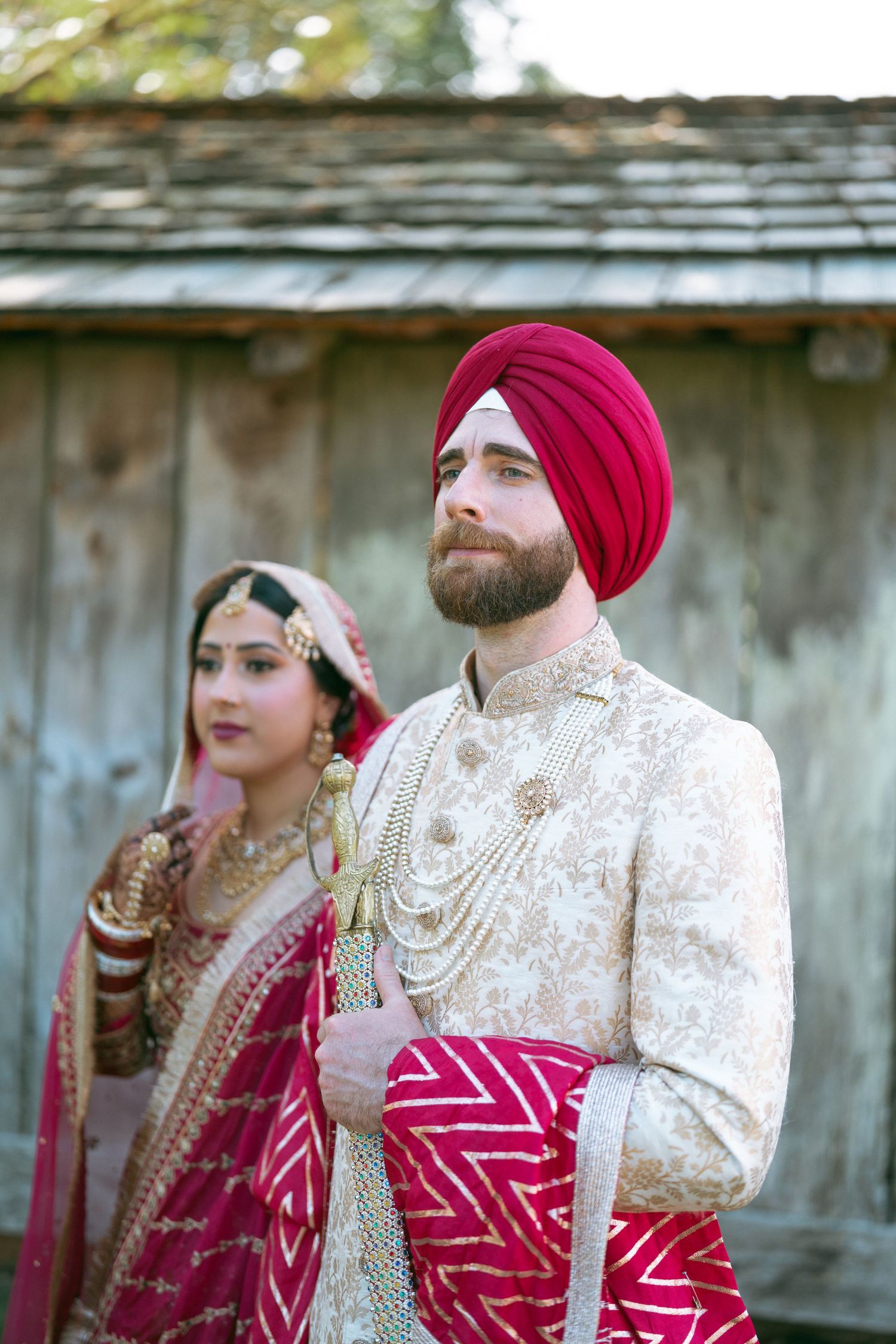 Sikh bride and groom in traditional red and gold wedding attire, standing outdoors near a rustic wooden structure.