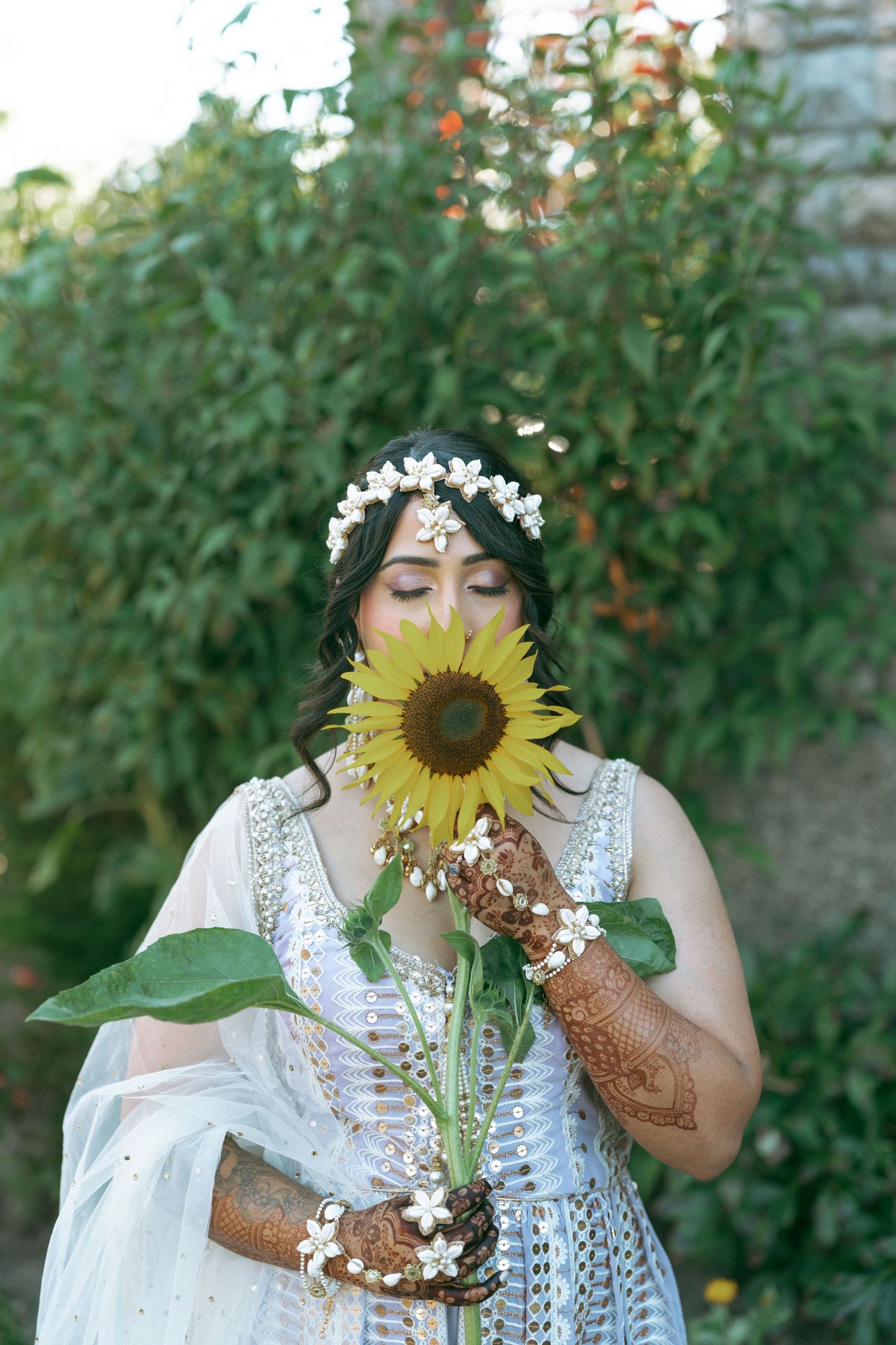 Bride in white dress with henna hands smells a sunflower, wearing a floral crown outdoors.